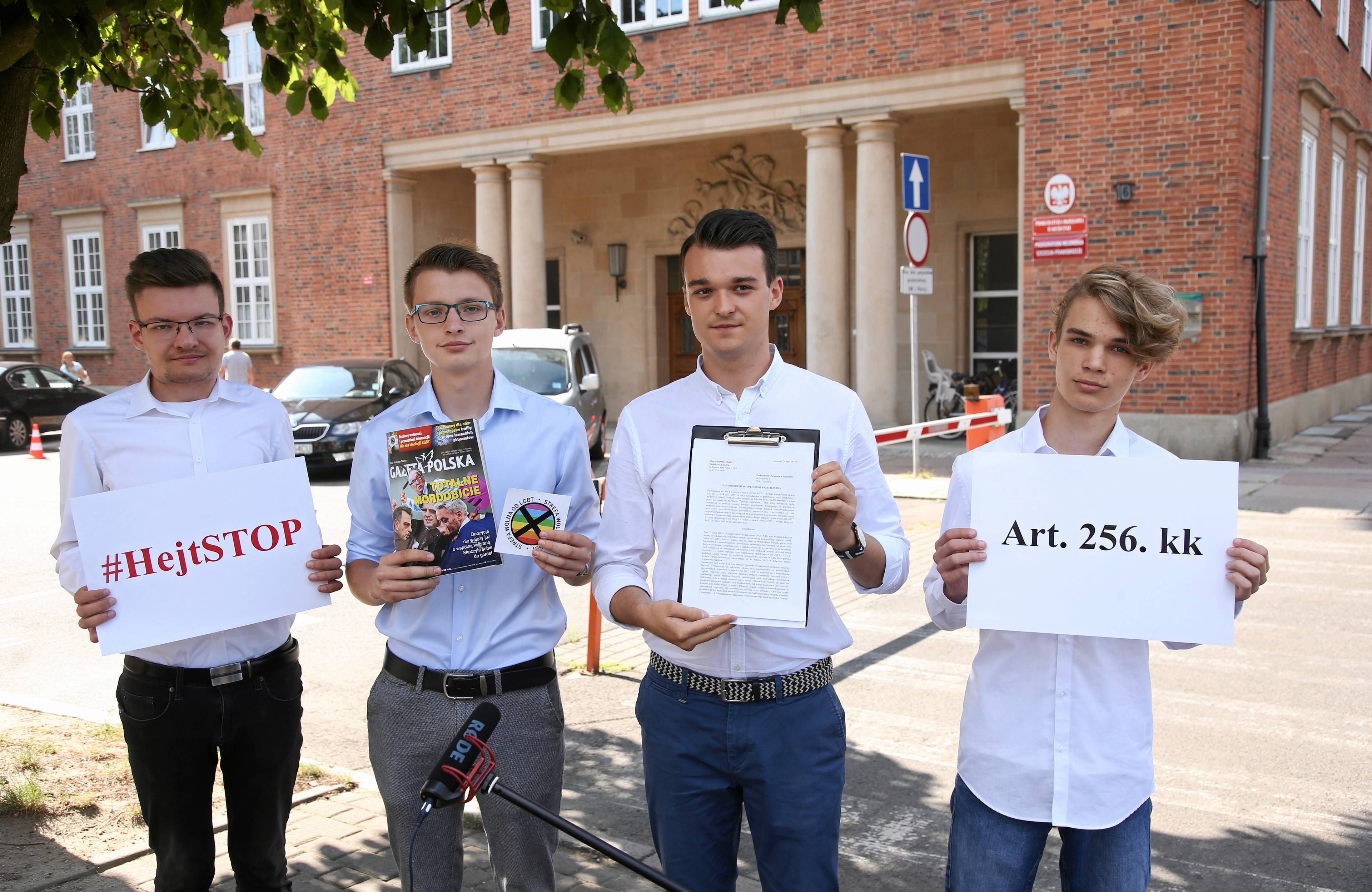 Czterech młodych mężczyzn w białych koszulach pozuje na tle budynku prokuratury w Szczecinie. Trzymają kartki z napisem "Hejt stop", "art. 256 kk" oraz wydanie Gazety Polskiej i dołączonej do niej naklejki w tęczowych kolorach z napisem "Strefa wolna od LGBT"
