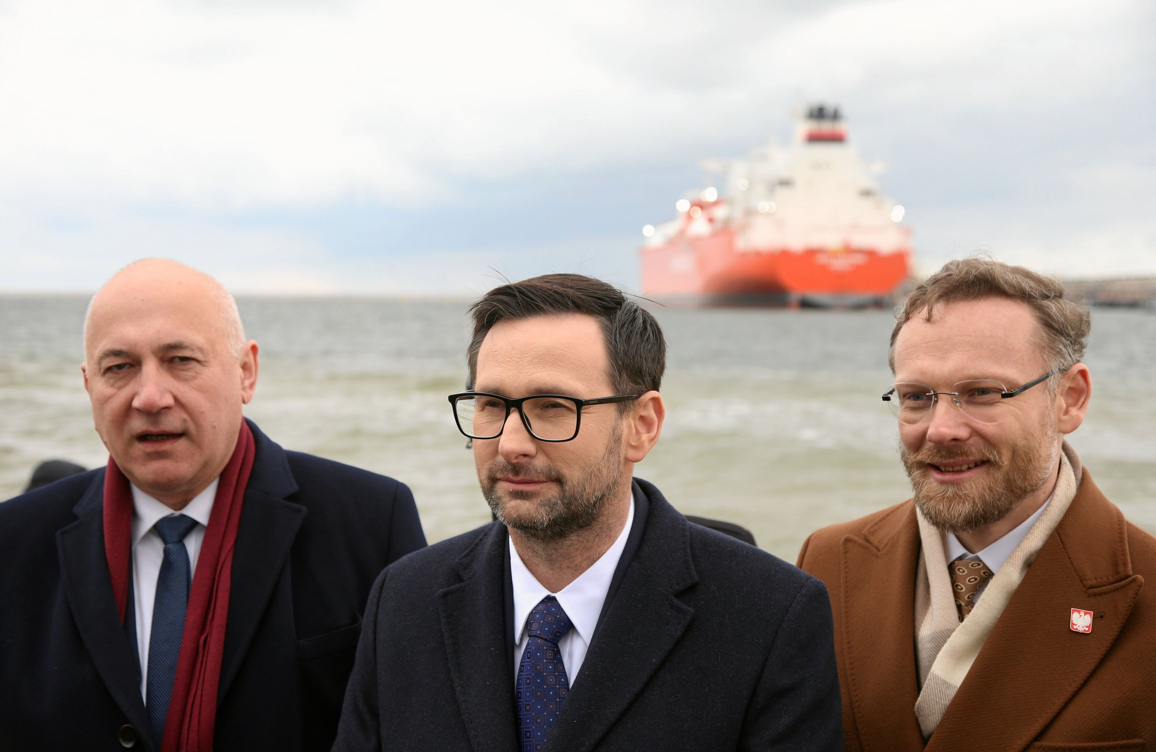 Trzech mężczyzn obok siebie nad wodą, za nimi biało-czerwony statek