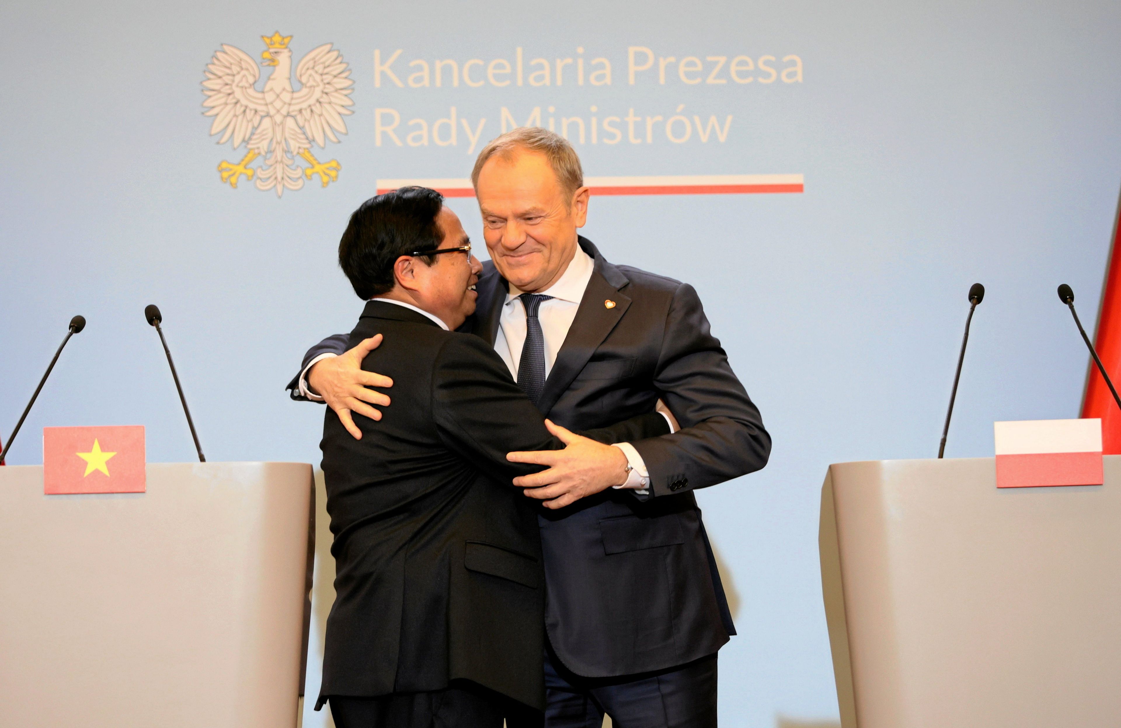 Dwaj mężczyźni, Europejczyk i Azjata, obejmują się przed ścianką z logo KPRM