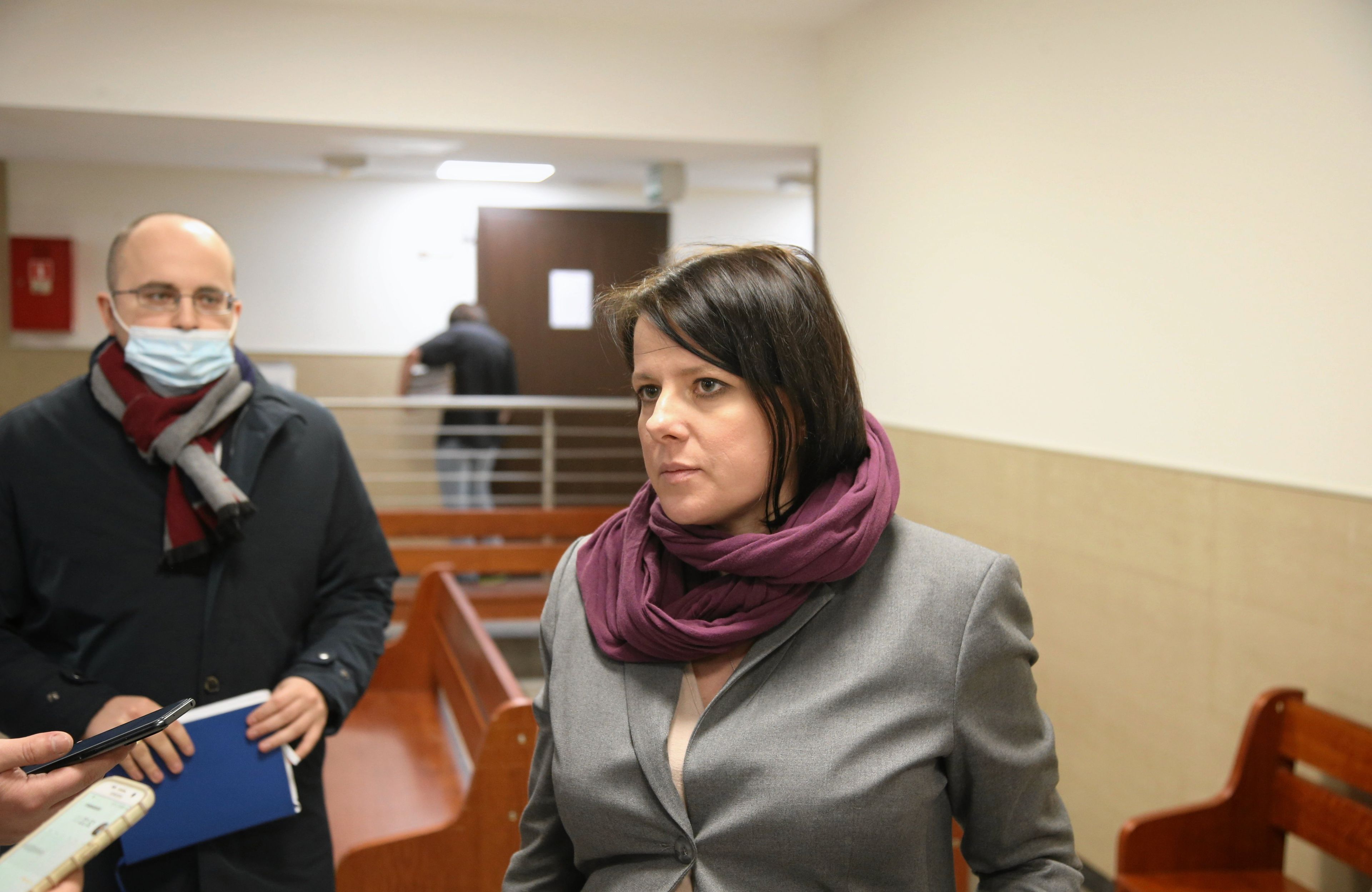 Kobieta z czarnymi włosami do ramion stoi przed mikrofonem, w tle mężczyzna w maseczce.