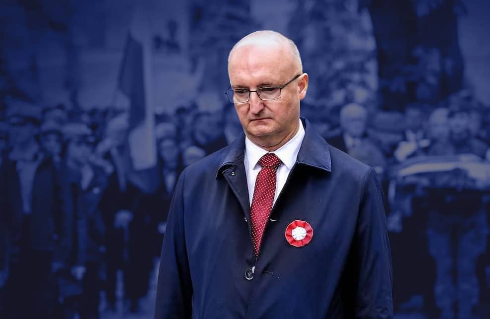 Wiceminister Piotr Wawrzyk na obchodach święta Konstytucji 3 Maja w Kielcach. Stoi ubrany w ciemny garnitur i płaszcz i spogląda ponuro w dół. W tle zgromadzenie ludzi z biało-czerwonymi flagami.