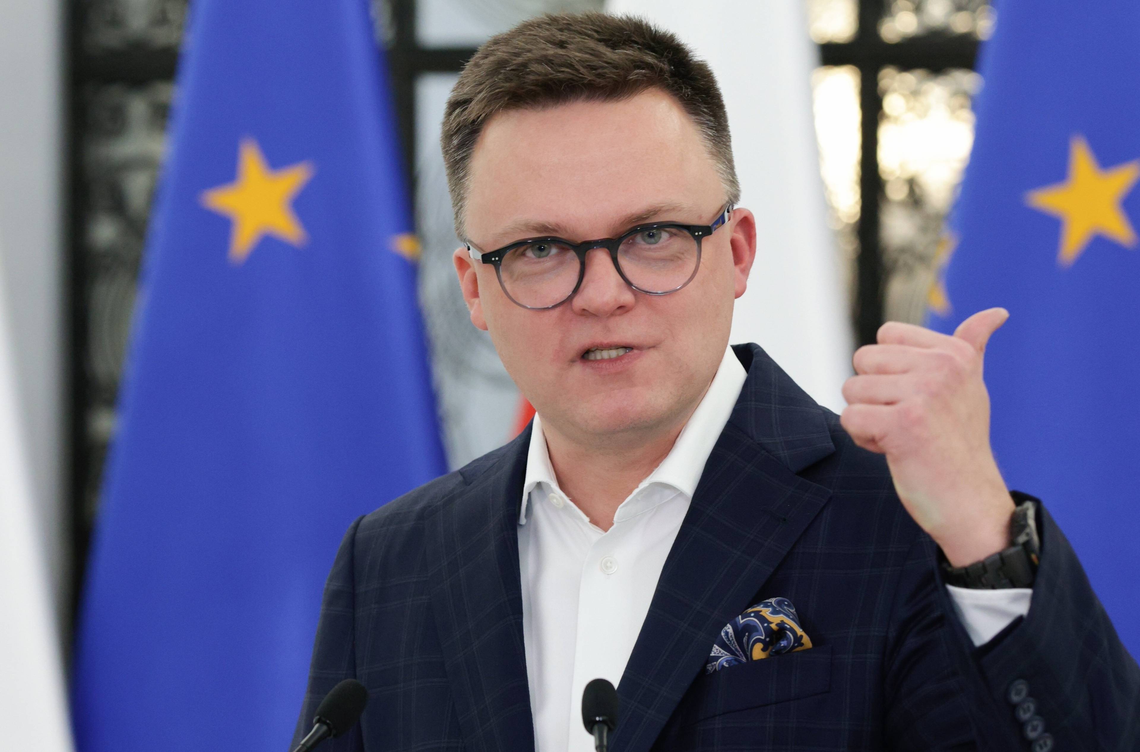 Ostrzyżony na jeża 40-latek w okularach, w garniturze bez krawata, na tle flag Polski i UE