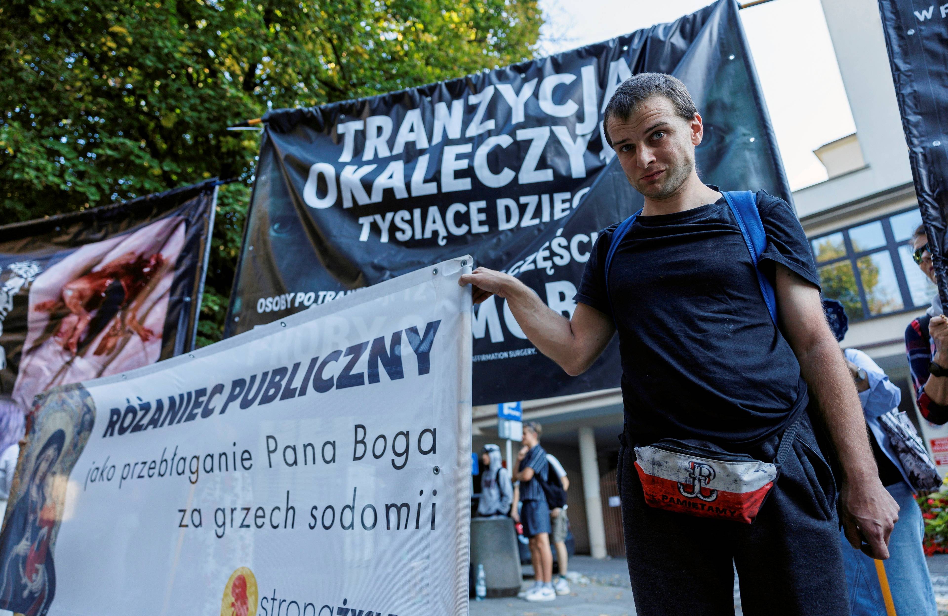 21.09.2024, Rzeszów. Kontrmanifestacja środowisk prawicowych na trasie V Marszu Równości w Rzeszowie. Młody mężczyzna ubrany w czarny t-shirt i biał-czerwoną nerkę ze znakiem Polski Walczącej trzyma transparent z napisem "Różaniec publiczny jak przebłaganie Pana Boga za grzech sodomii". Za nim transparent z napisem "Tranzycja okaleczyła tysiące dzieci"