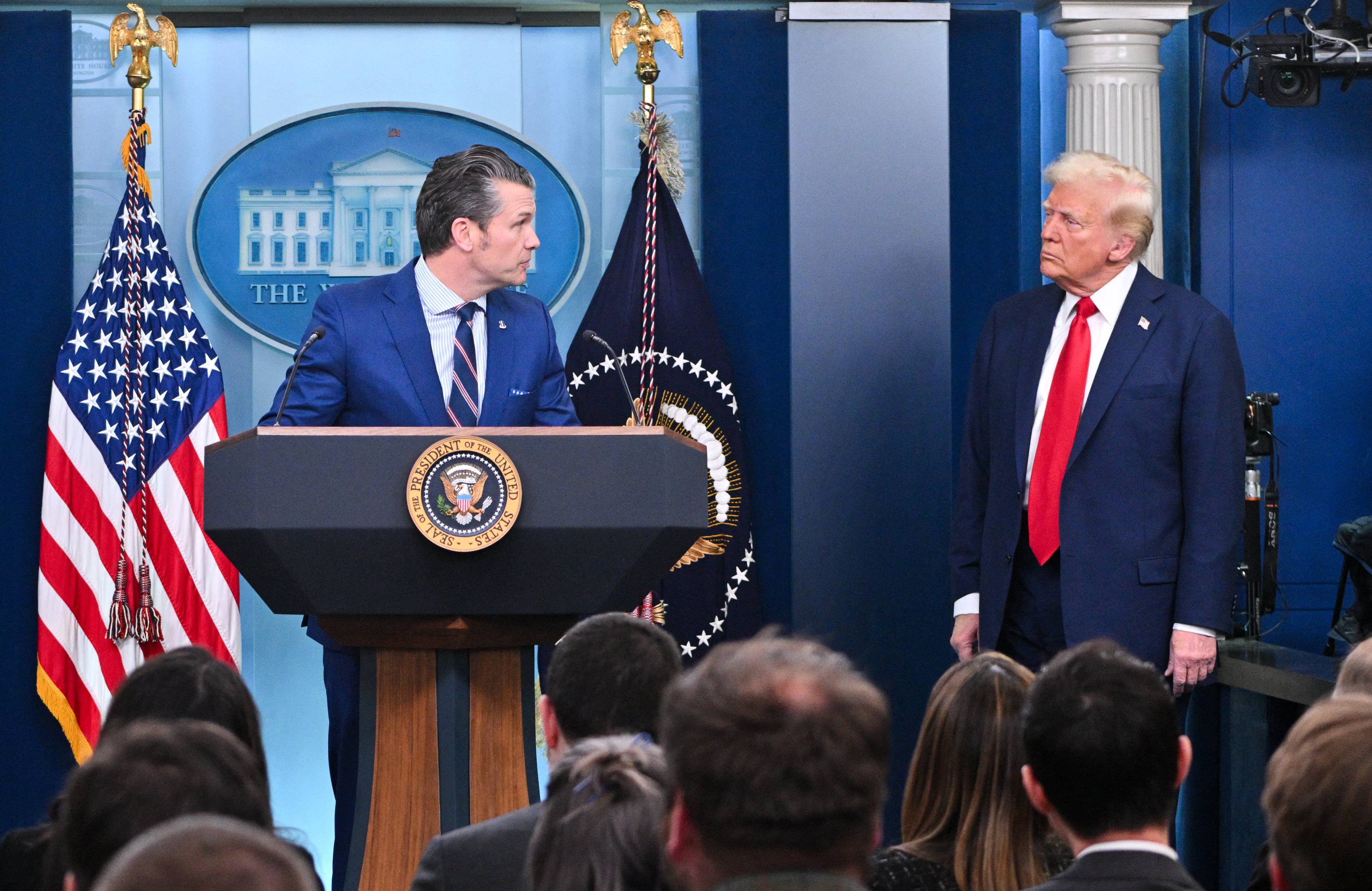Na zdjęciu widzimy Donalda Trumpa w granatowym garniturze i czerwonym krawacie, prezydent USA stoi z boku podestu. Na środku przy mównicy stoi Pete Hegseth sekretarz obrony, wysoki mężczyzna w niebieskim garniturze, z siwymi włosami. Konferencja odbywa się w Białym Domu.