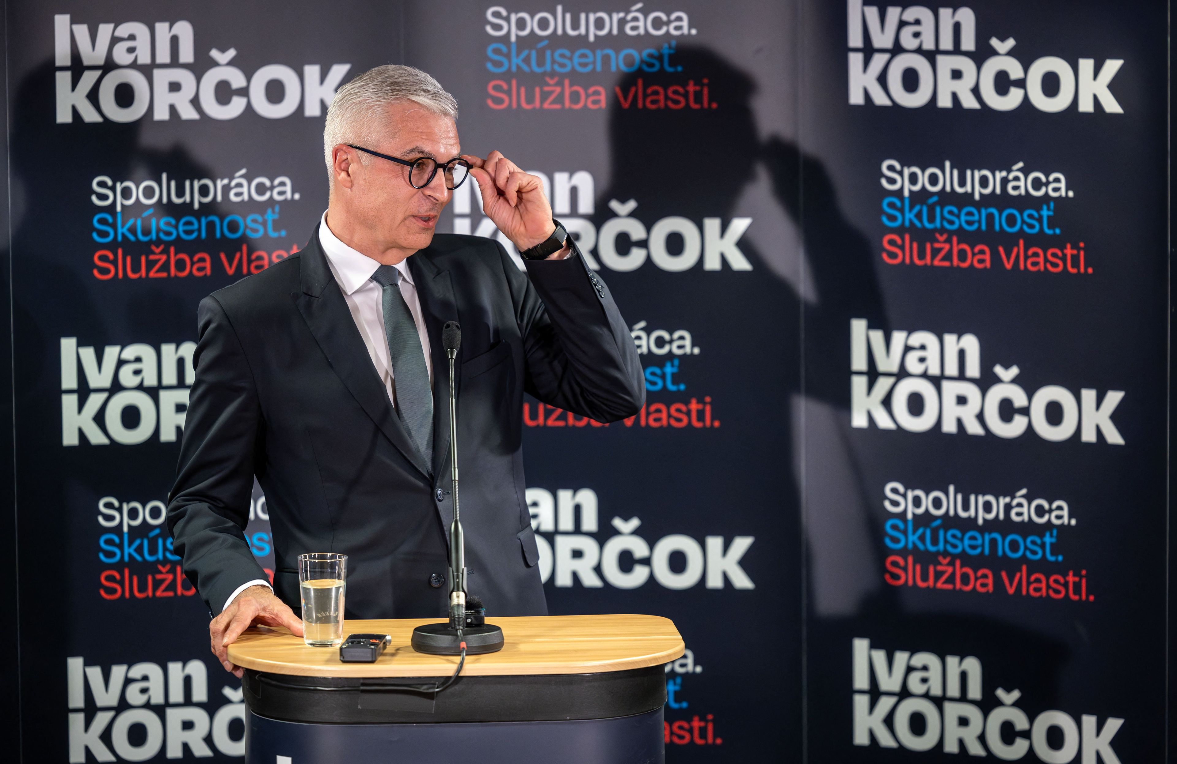 Męzczyzna przemawia na tle plakatów wyborczych z nazwiskiem "Ivan Korcok"
