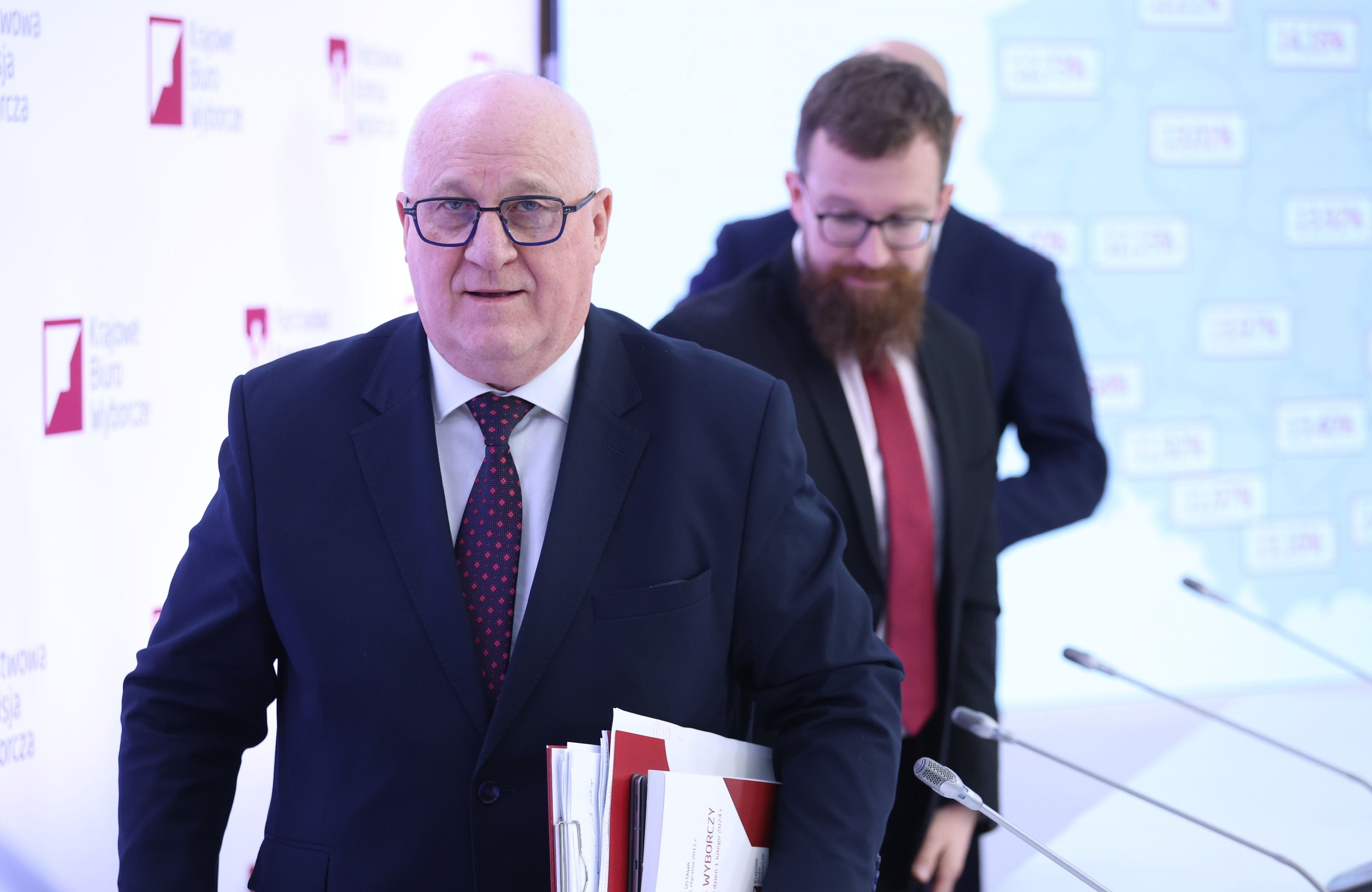 Korpulentny łysy mężczyzna w garniturze i okularach, za nim młodszy z brodą i wąsami.