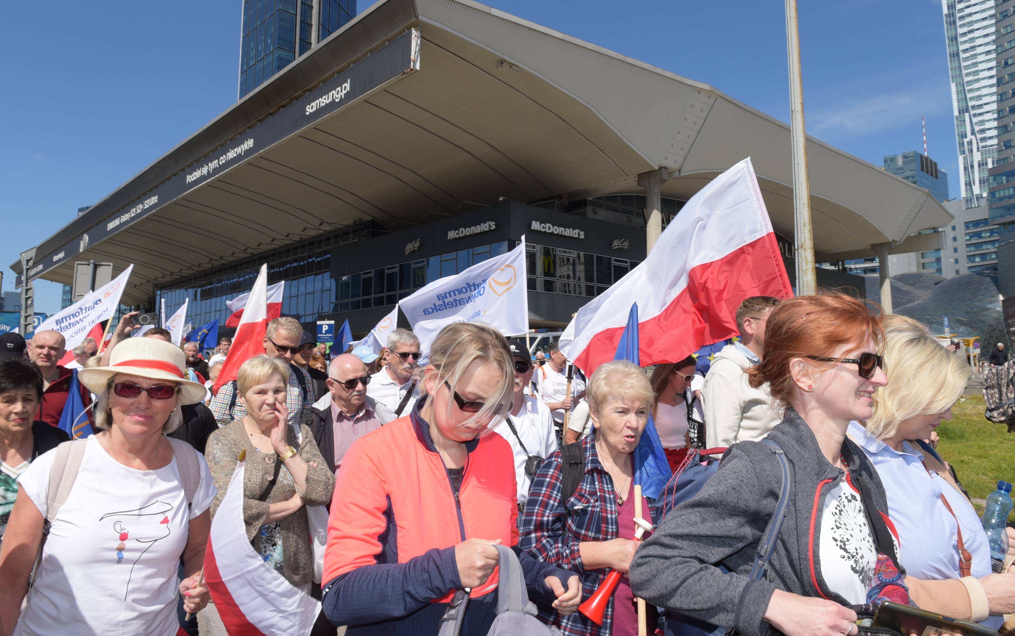 Ludzie z biało-czerwonymi flagami przed Dworcem Centralnym w Warszawie, w tle szklane biurowce