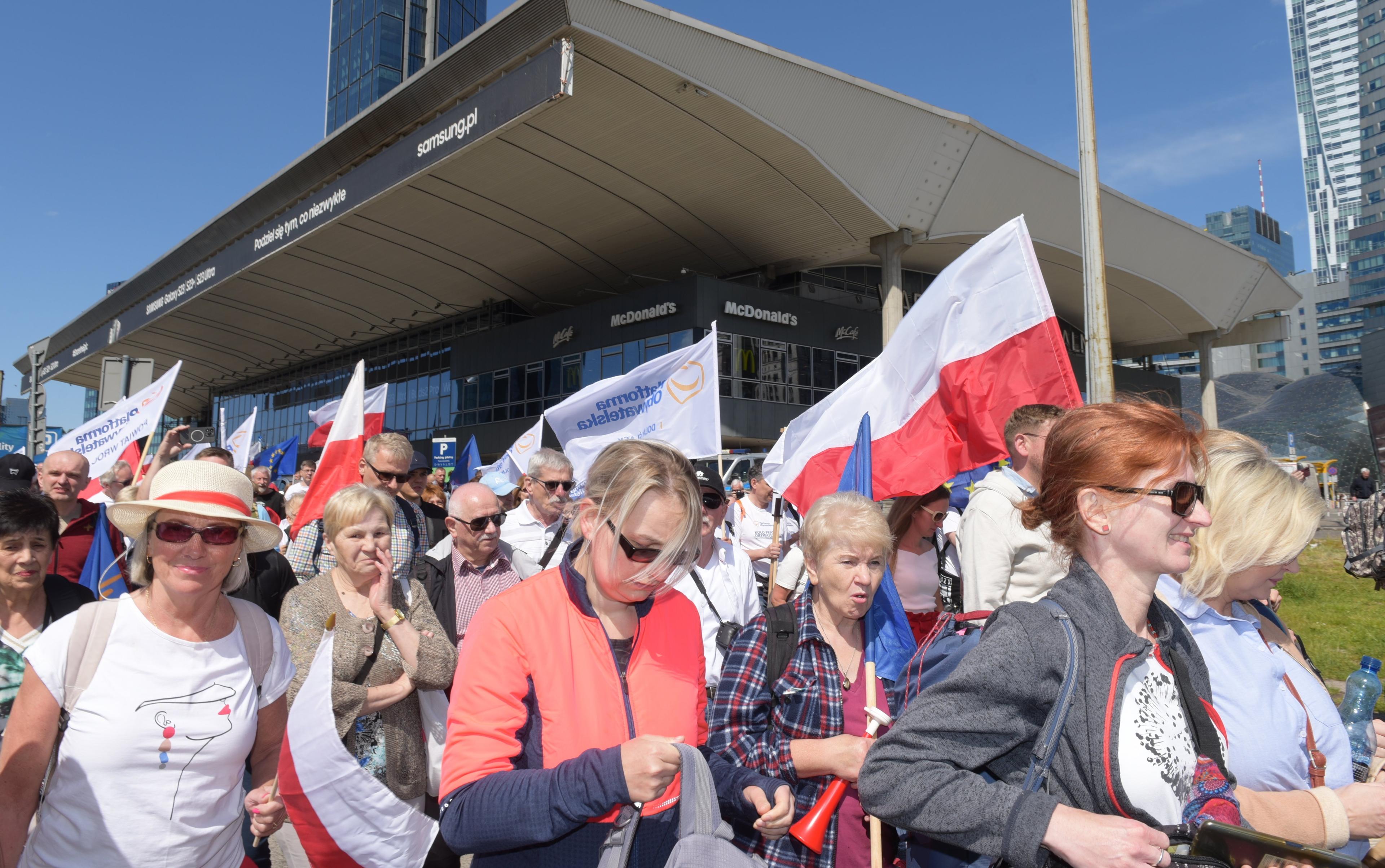 Ludzie z biało-czerwonymi flagami przed Dworcem Centralnym w Warszawie, w tle szklane biurowce