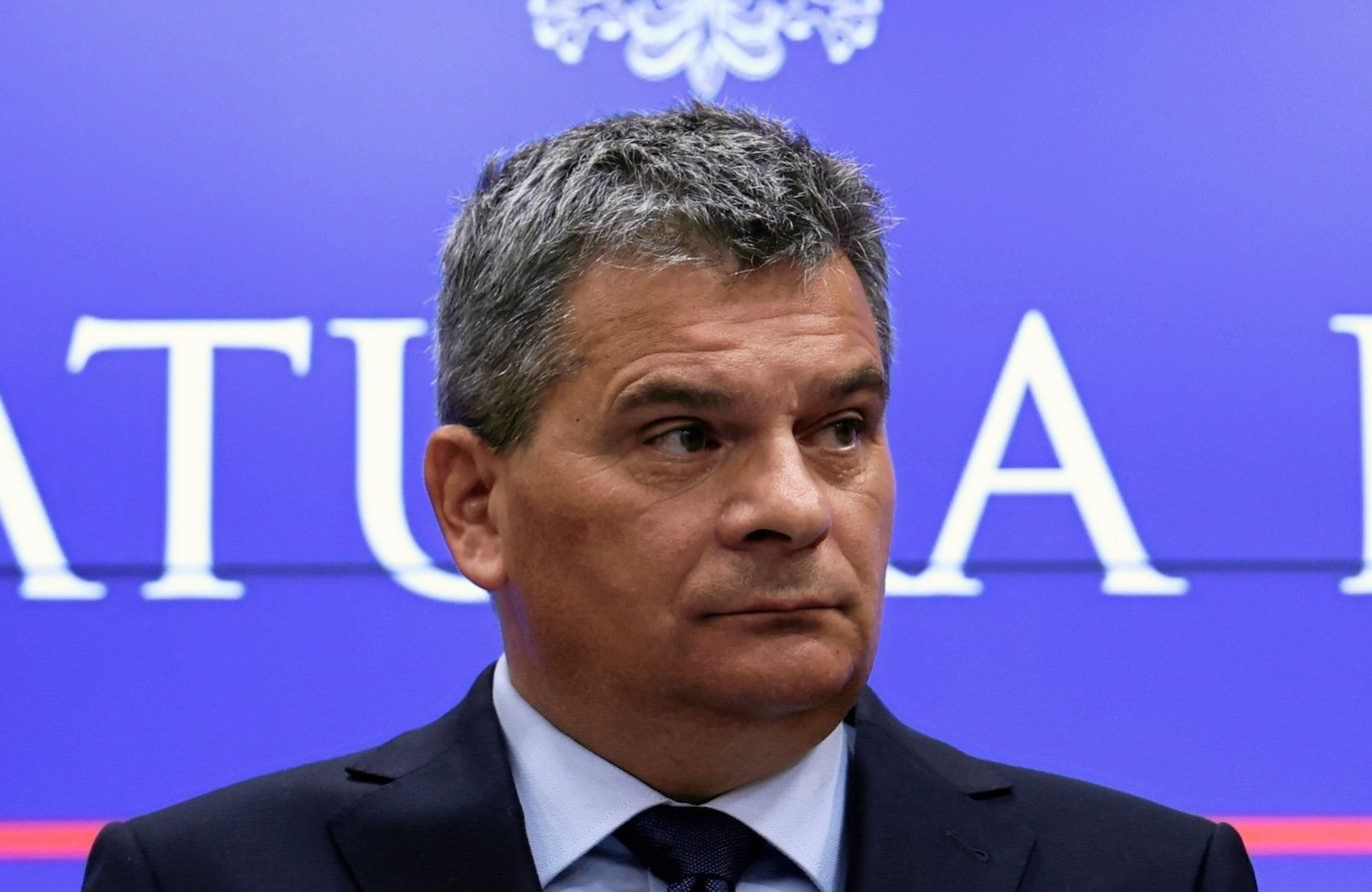 Prokurator Krajowy Dariusz Korneluk w siedzibie prokuratury, na tle ścianki z napisem prokuratura krajowa. Nad głową ma orzełka – polskie godło.