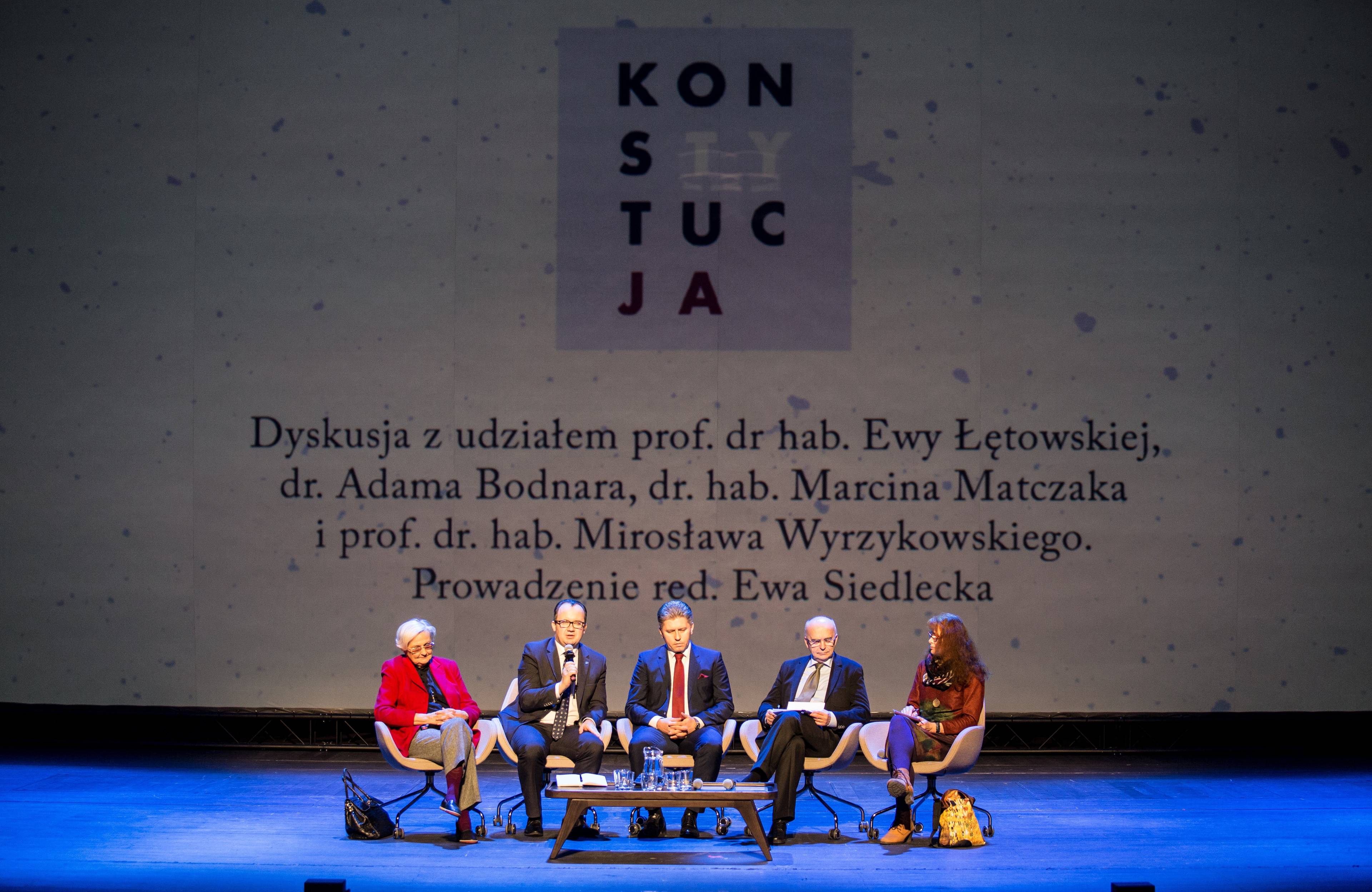 Pięć osób siedzi na scenie podczas dyskusji. W tle na ekranie projekcja przedstawiająca plakat „KonstytucJA” i lisą nazwisk dyskutantów