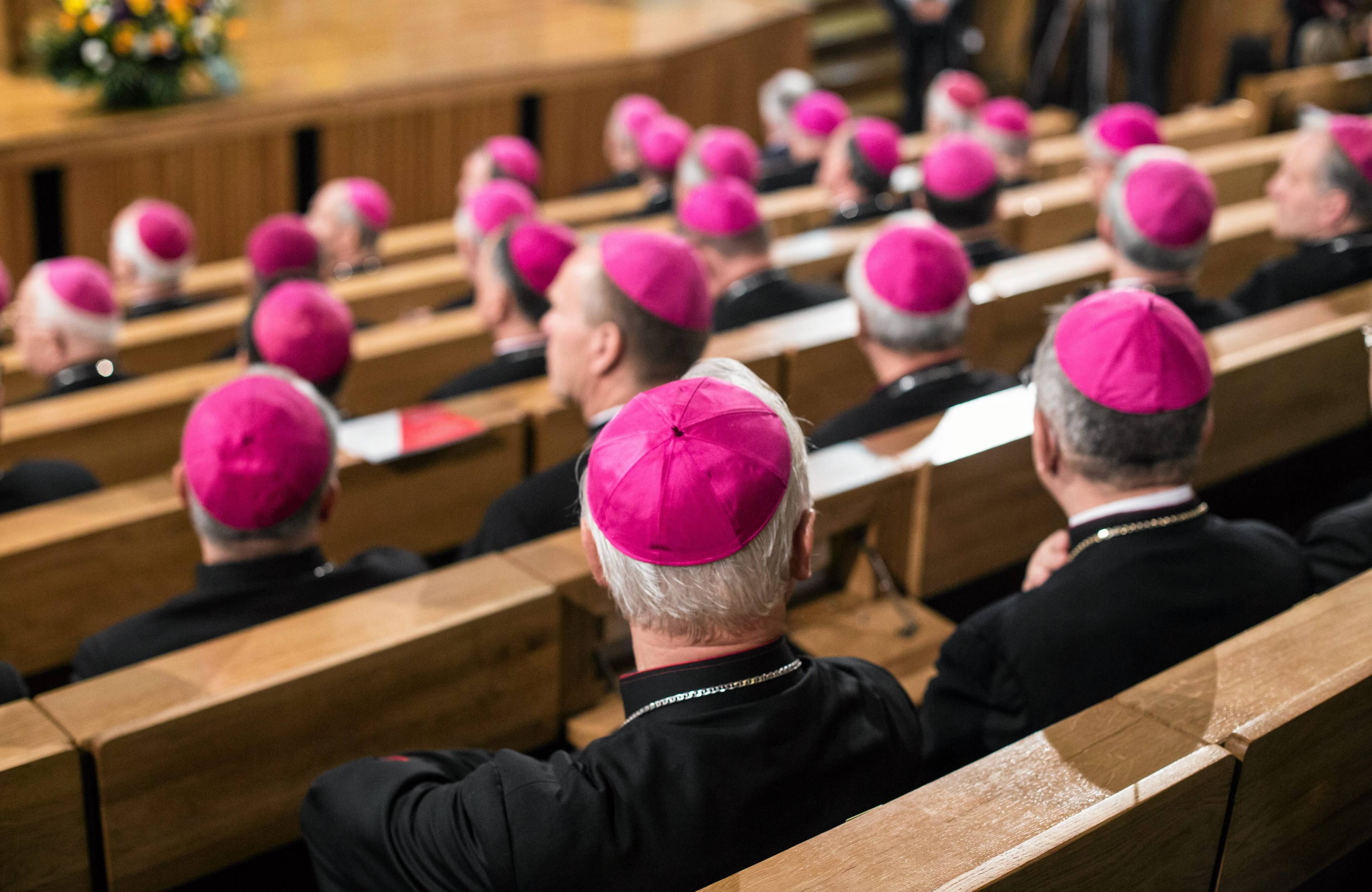 Mężczyźni w purpurowych piuskach siedzący w ławach audytorium, widziani od tyłu. Kościół