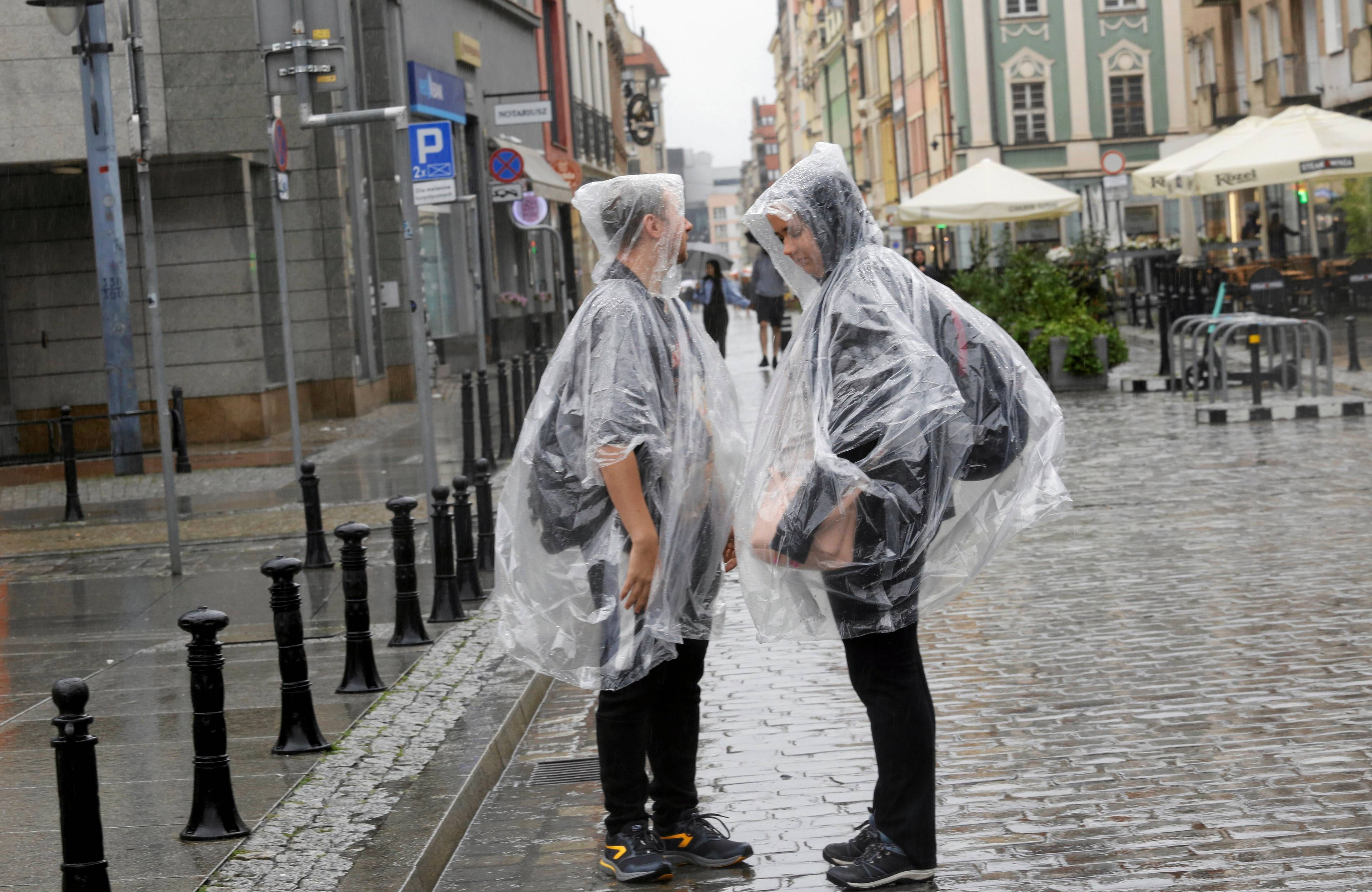 Wrocław, 06.08.2023. Dwóch młodych mężczyzn w pelerynach przeciwdeszczowych stoi w deszczu na wrocławskiej ulicy.