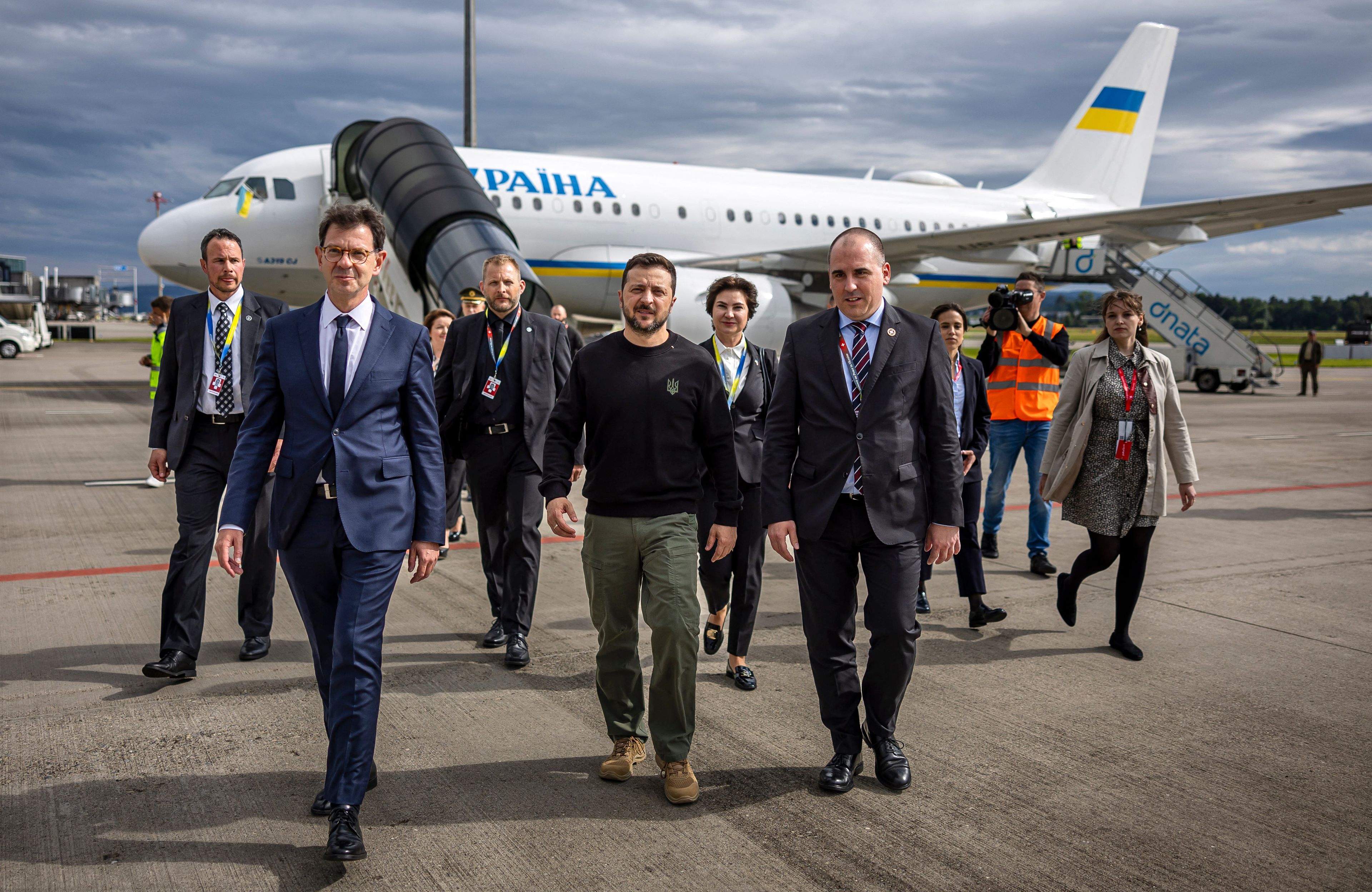 Na zdjęciu jest grupa osób, które idą. Po środku jest prezydent Ukrainy Wołodymyr Zełenski. W tle jest biały samolot z napisam "Ukraina".