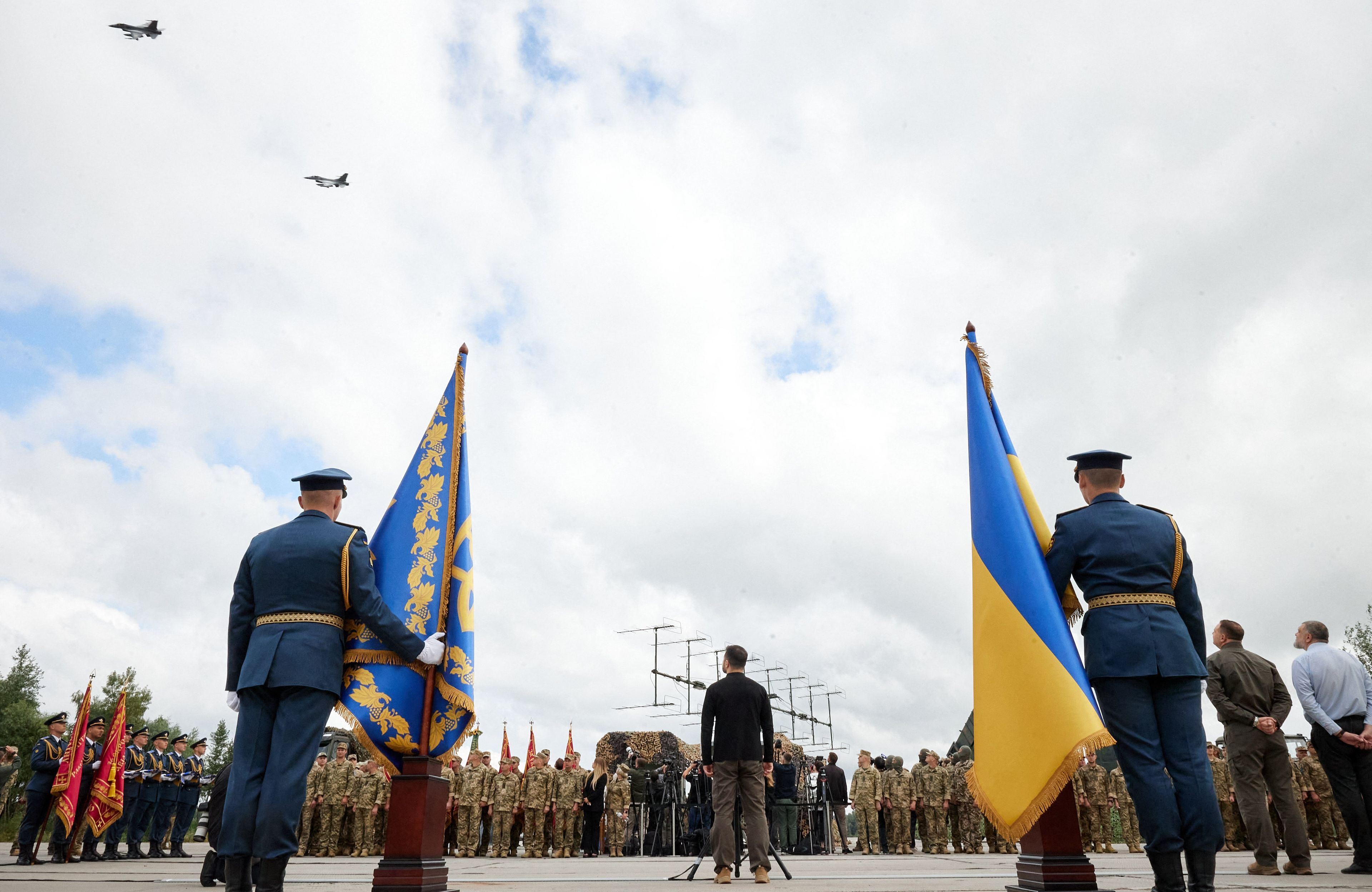 Na zdjęciu widać trzech mężczyn, stojących tyłem. Dwóch jest ubranych w granatowe mundury, trzymają flagi w niebiesko-żólntych kolorach: flagę z godłem Ukrainy oraz flagę Ukrainy. Pośrodku jest mężczyzna ubrany w czarną bluzę i spodnie koloru khaki. W tle widać mężczyzn w wojskowych mundurach polowych. W niebie widać dwa myśliwce.