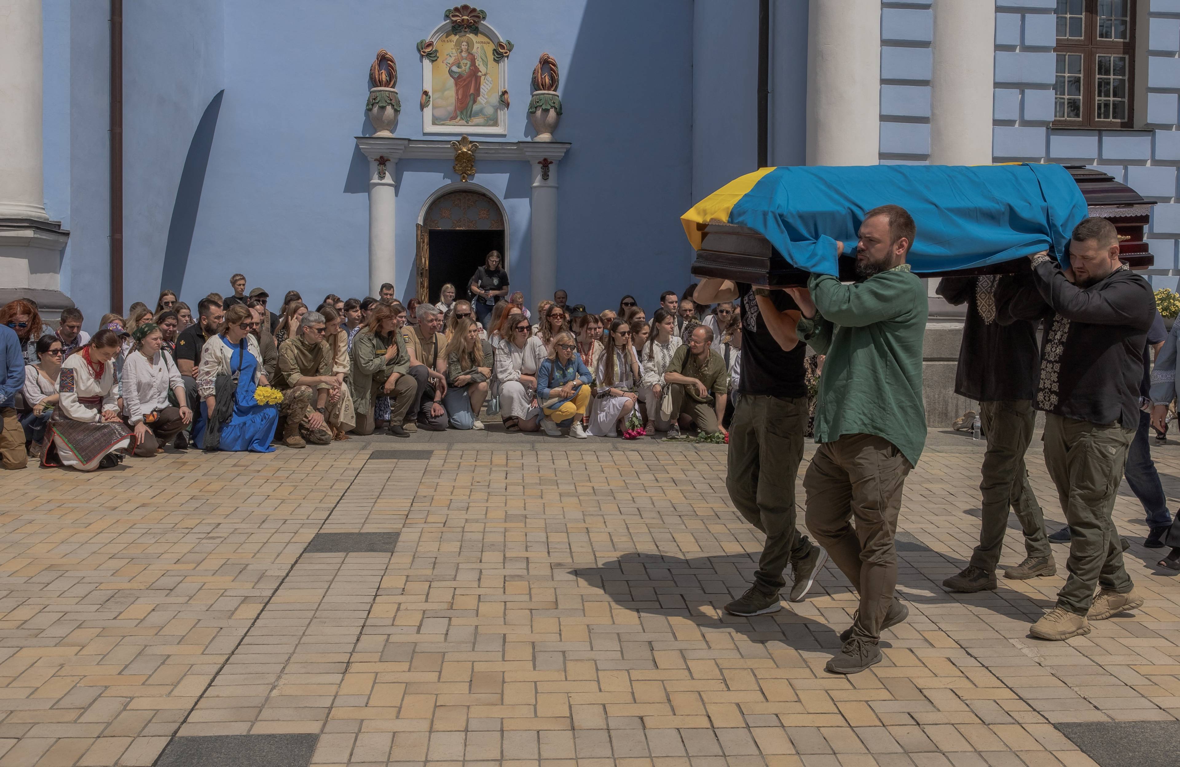 mężczyźni niosą trumnę spowitą w niebiesko-zółtą flagę, w tle cerkiew i klęczący żałobnicy