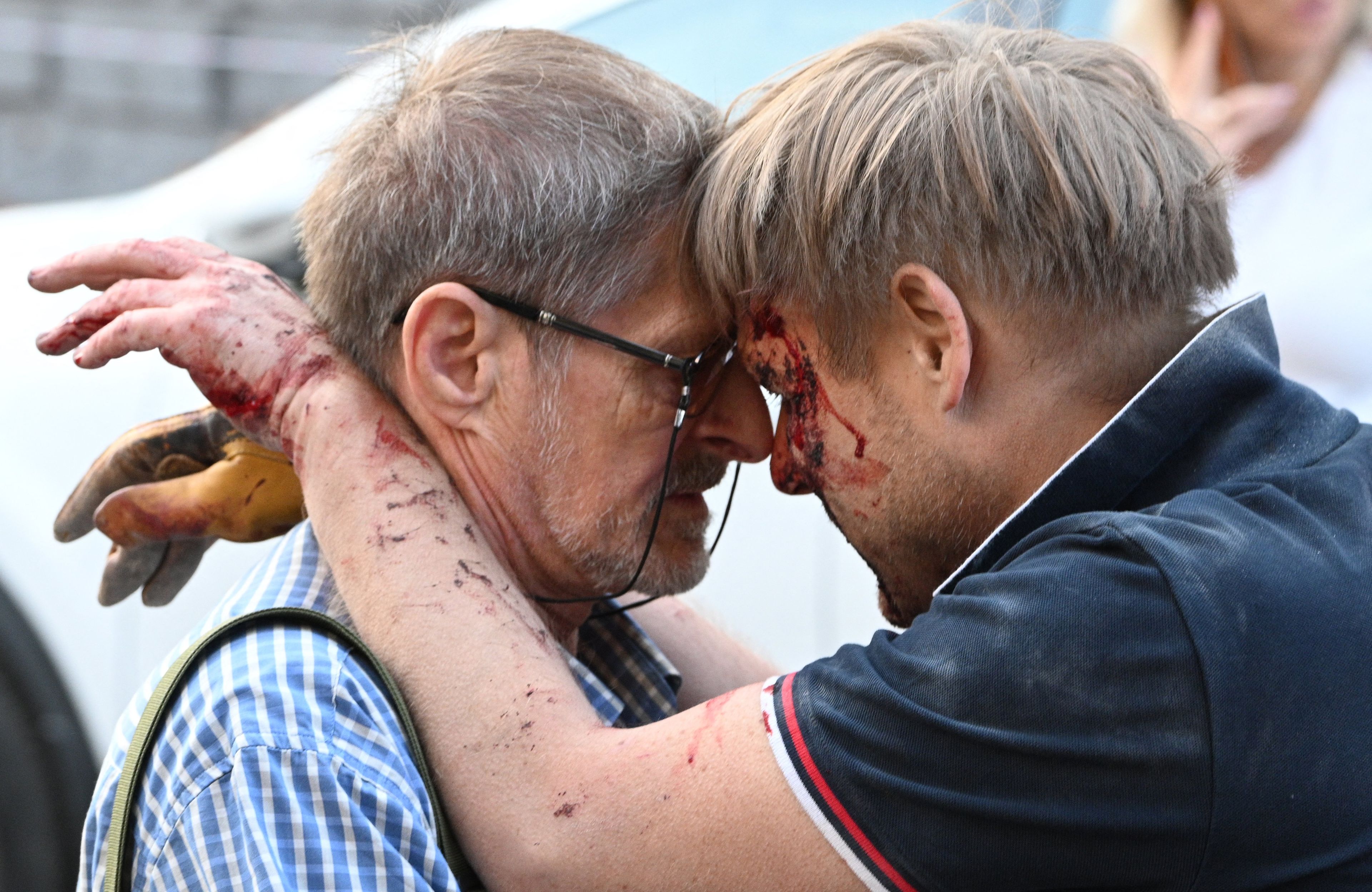Na zdjęciu widać dwóch mężczyzn. Mężczyzna po prawej opiera się o starszego mężczyznę po lwej. Jest w czarnej koszuli, ma zakrwawioną twarz i ręce.
