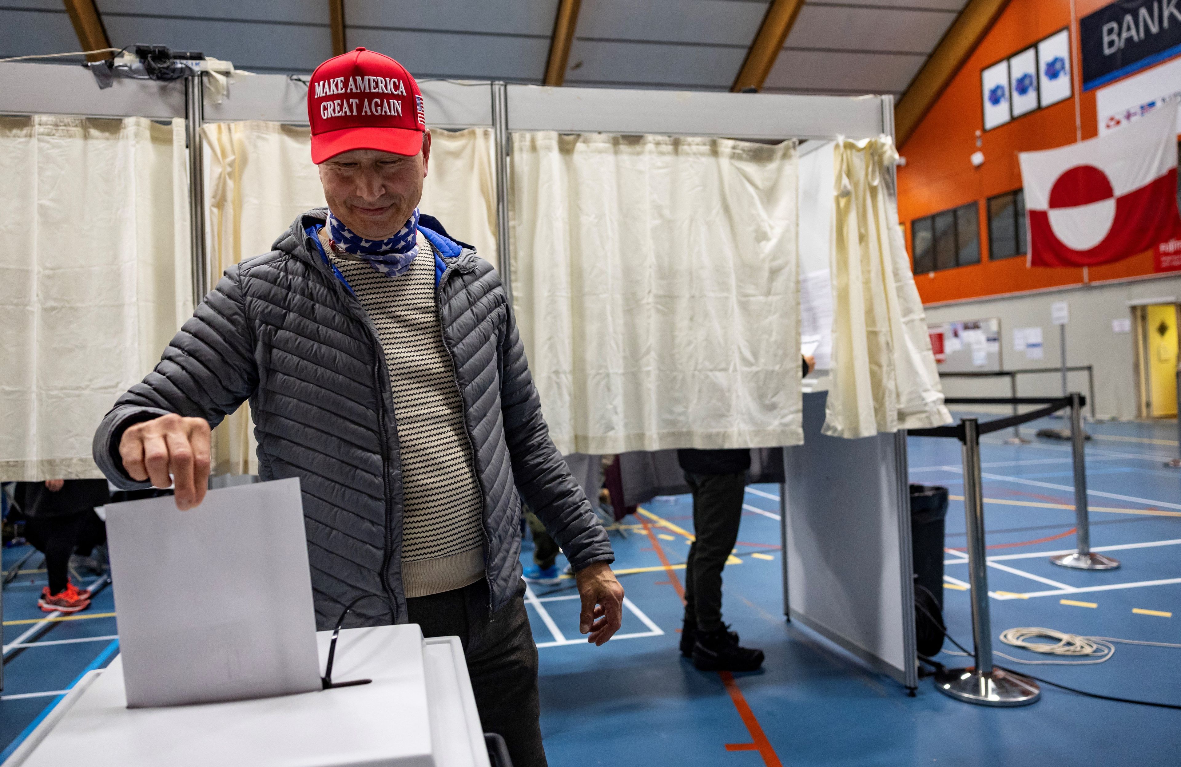 Mężczyzna w czerwonej czapce z daszkiem z napisem "Make America Great Again" i z chustką na szyi w kolorze amerykańskiej flagi wrzuca głos do urny wyborczej. Za nim na ścianie lokalu wyborczego wisi biało-czerwona flaga Grenlandii