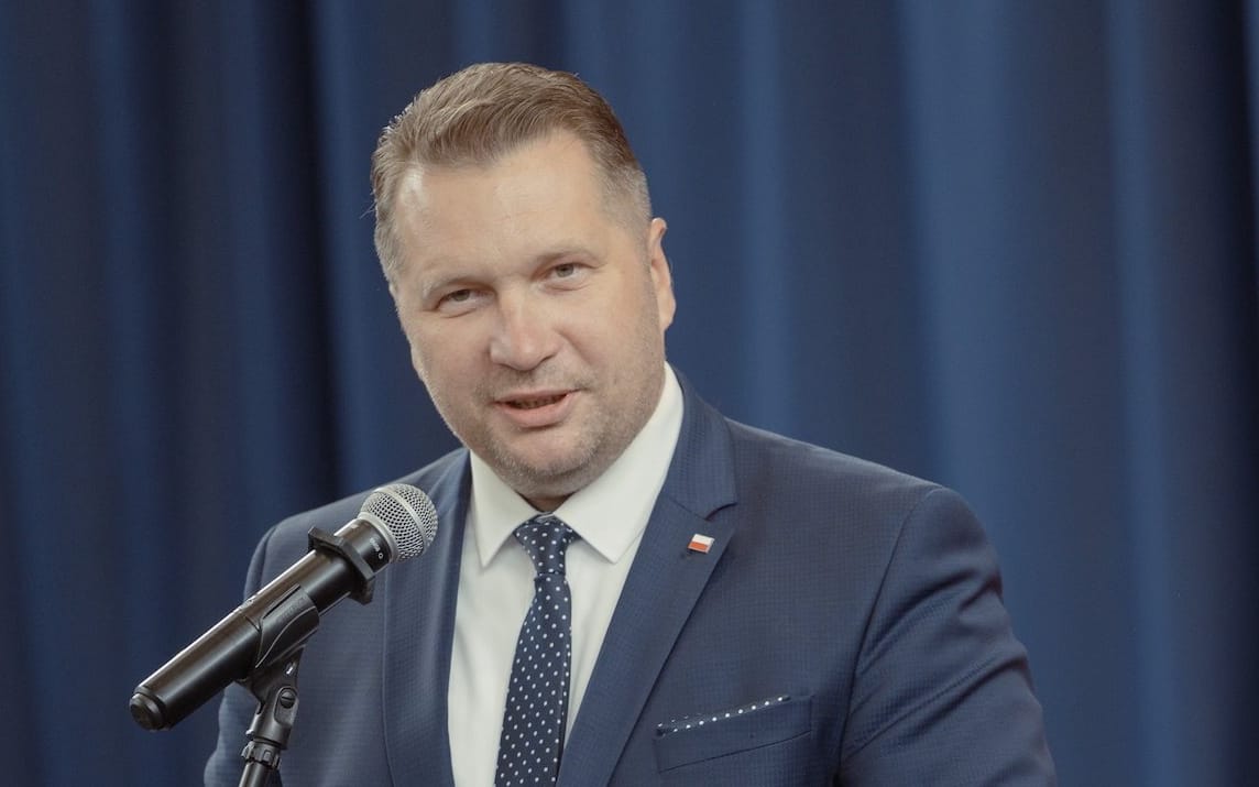 Minister Edukacji Przemysław Czarnek przemawia do mikrofonu ubrany w szaroniebieski garnitur i granatowy krawat w białe kropki.