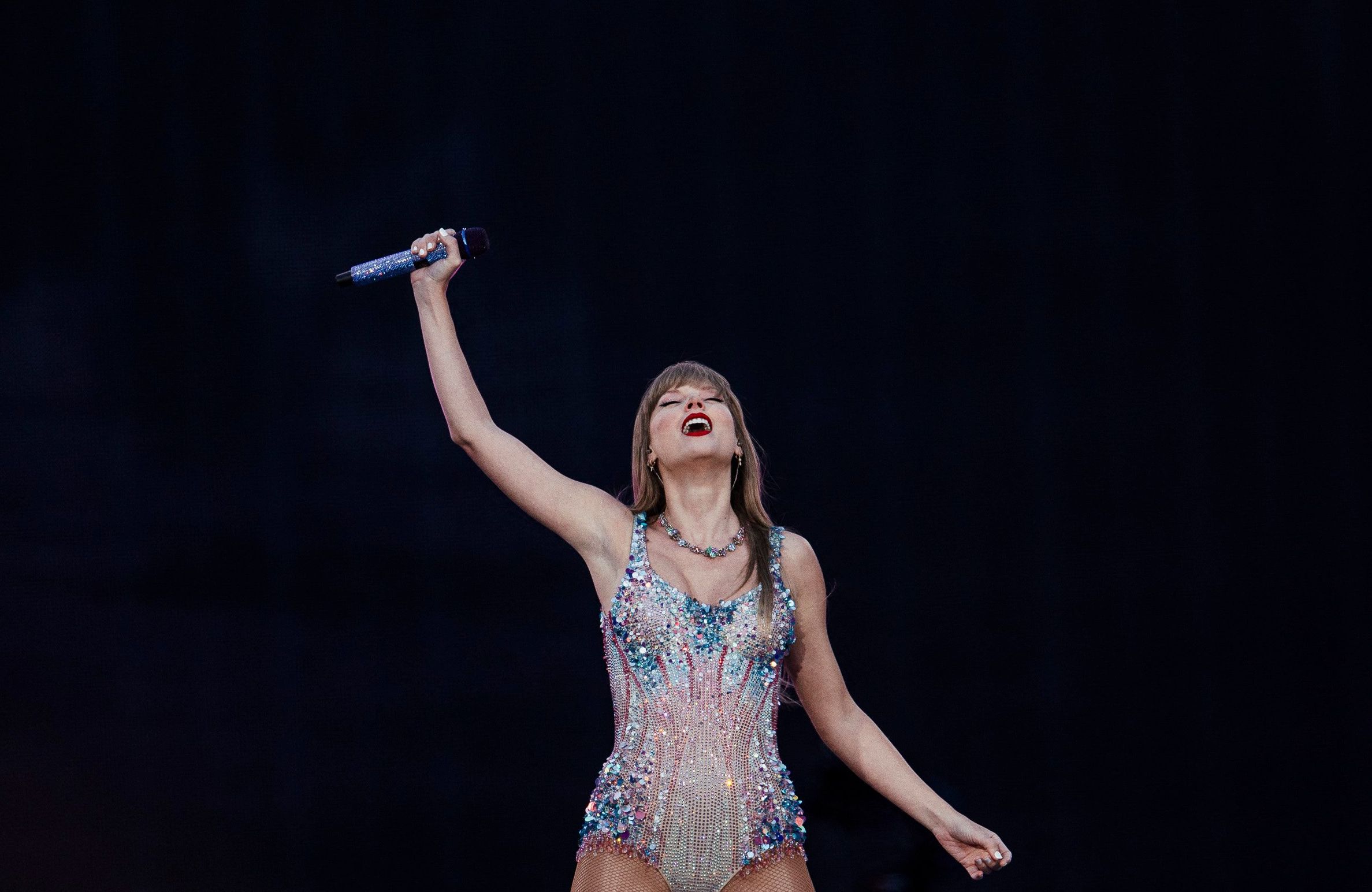 Piosenkarka Taylor Swift w jaskrawym kostiumie stoi na scenie z głową odchyloną do tyłu i prawą ręką wzniesioną w górę