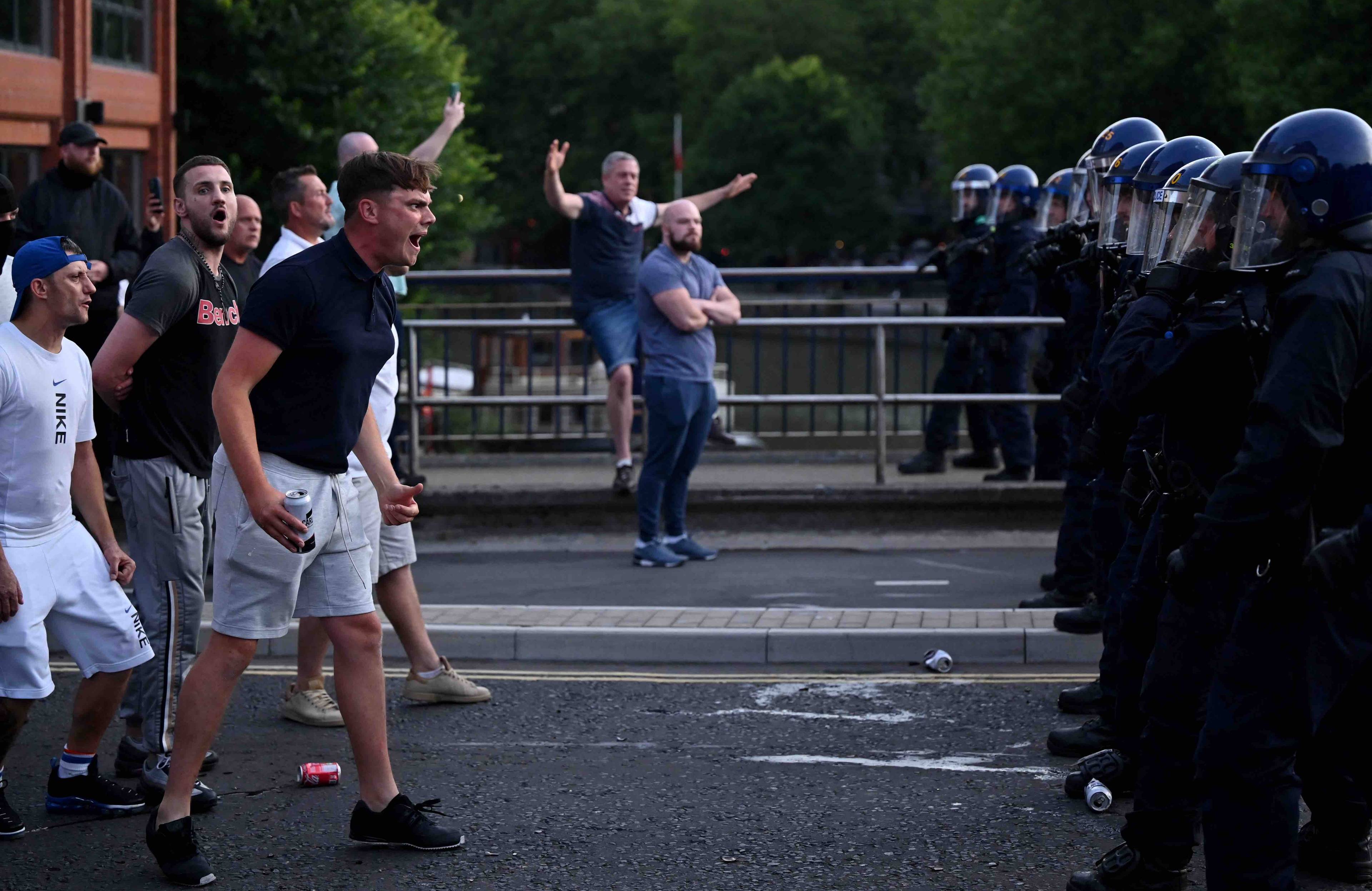 Młodzi mężczyźni stoją w poprzek ulicy i krzyczą w stronę szeregu policjantów w hełmach