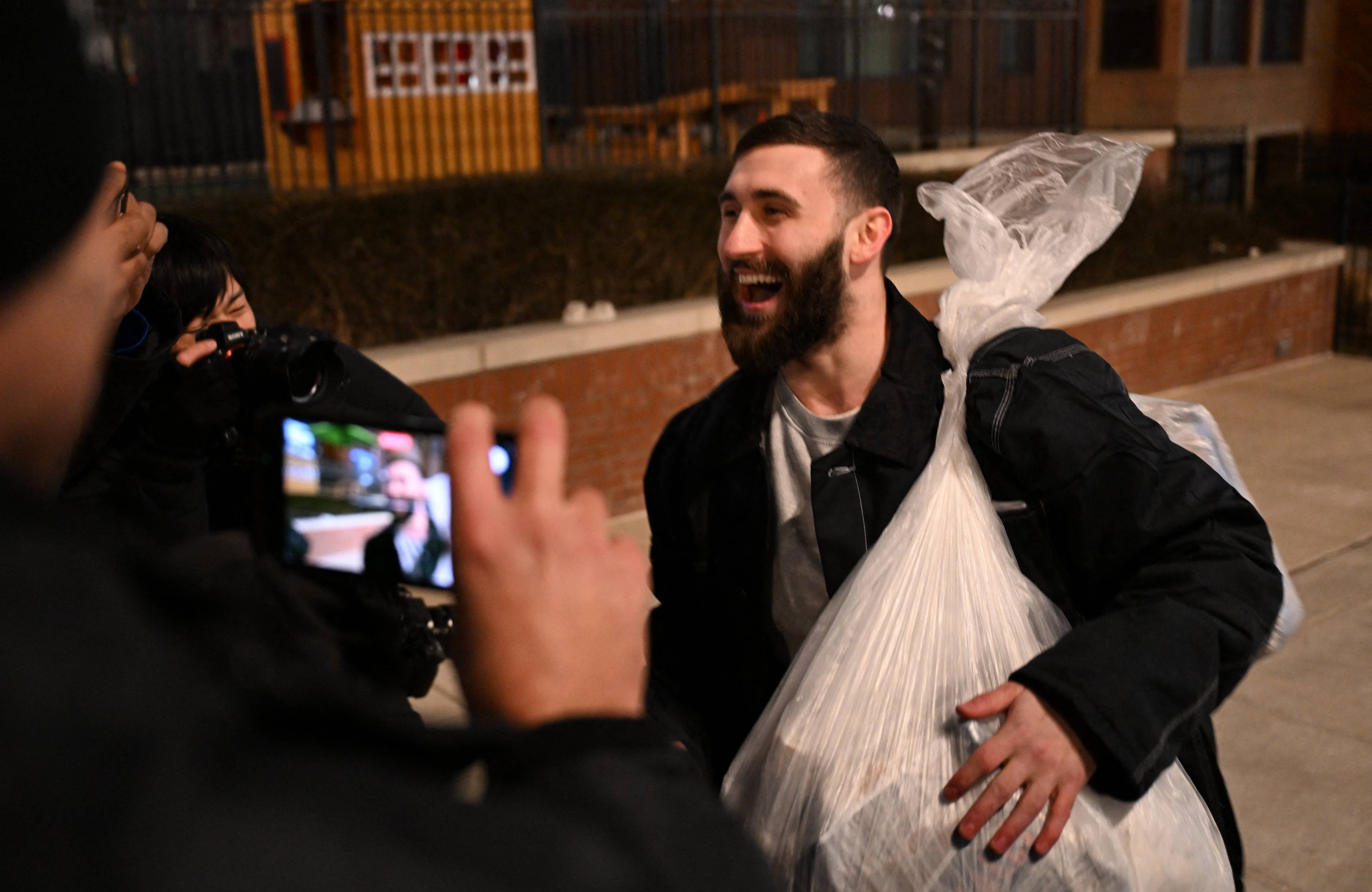 Mężczyzna z czarną brodą i plastikowym workiem na ramieniu wita się radośnie z postaciami stojącymi tyłem na pierwszym planie.