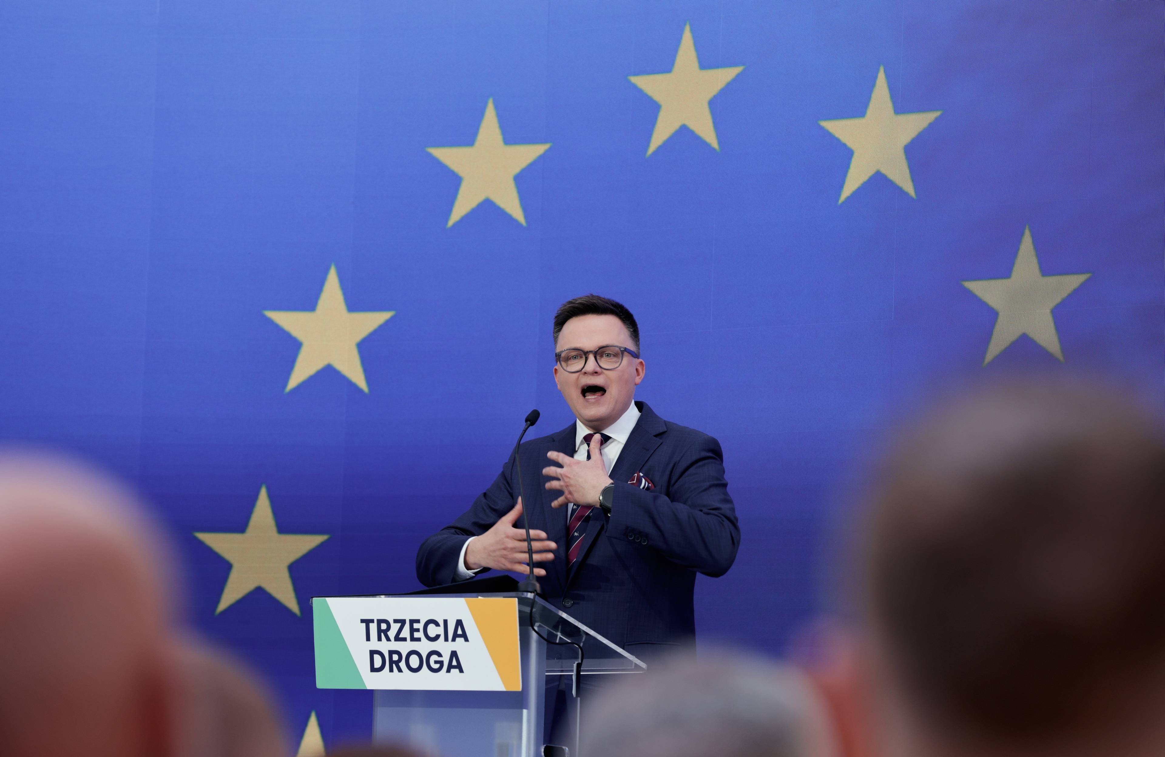 Szymon Hołownia przemawia na konwencji przed eurowyborami. Stoi za mównicą z napisem "Trzecia Droga", za nim niebieskie tło i gwiazdki z flagi Unii Europejskiej. Wybory europejskie