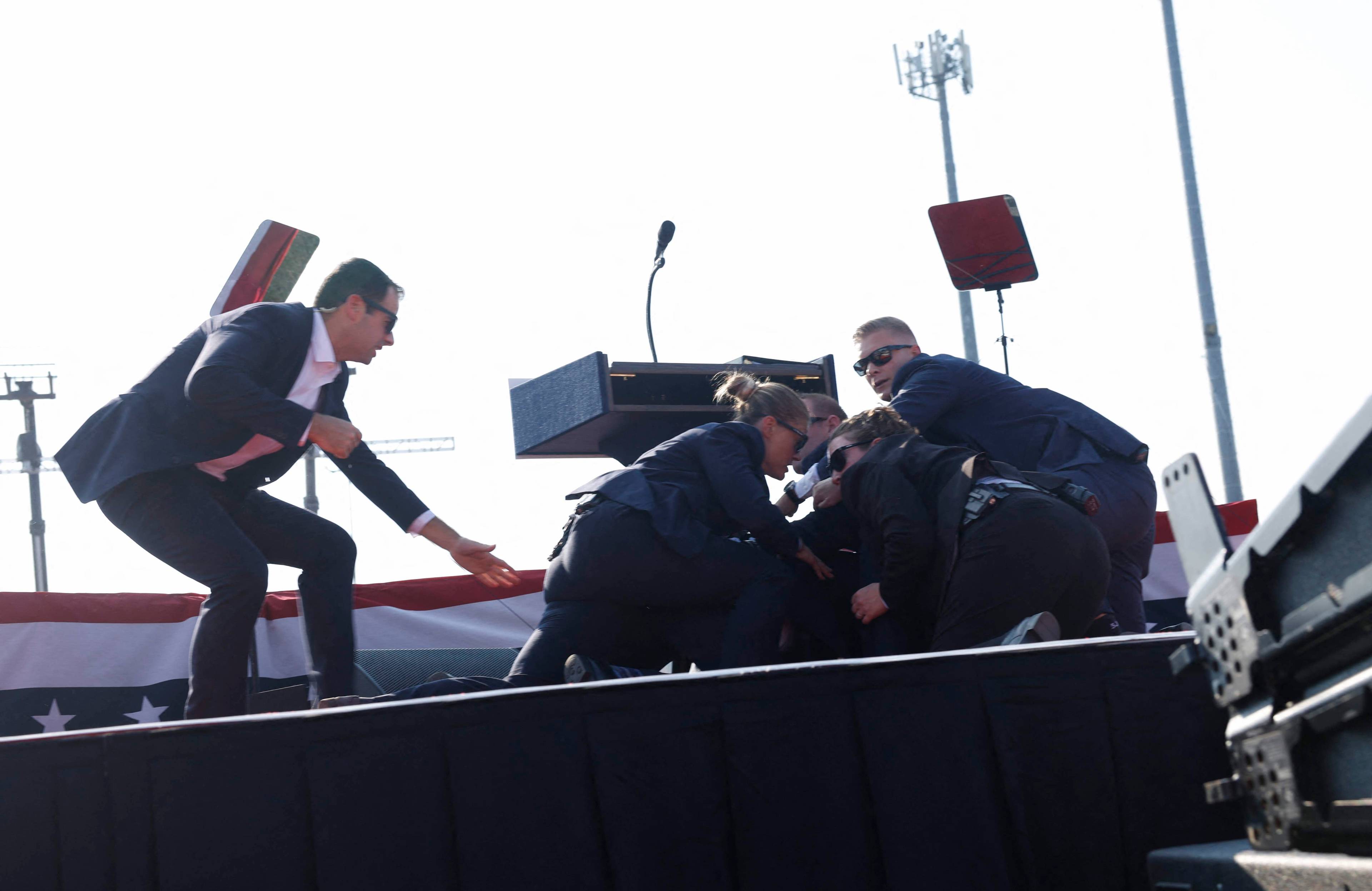 Agenci ochrony przykrywają swoimi ciałami Donalda Trumpa, leżącego na scenie. Zamach na Trumpa