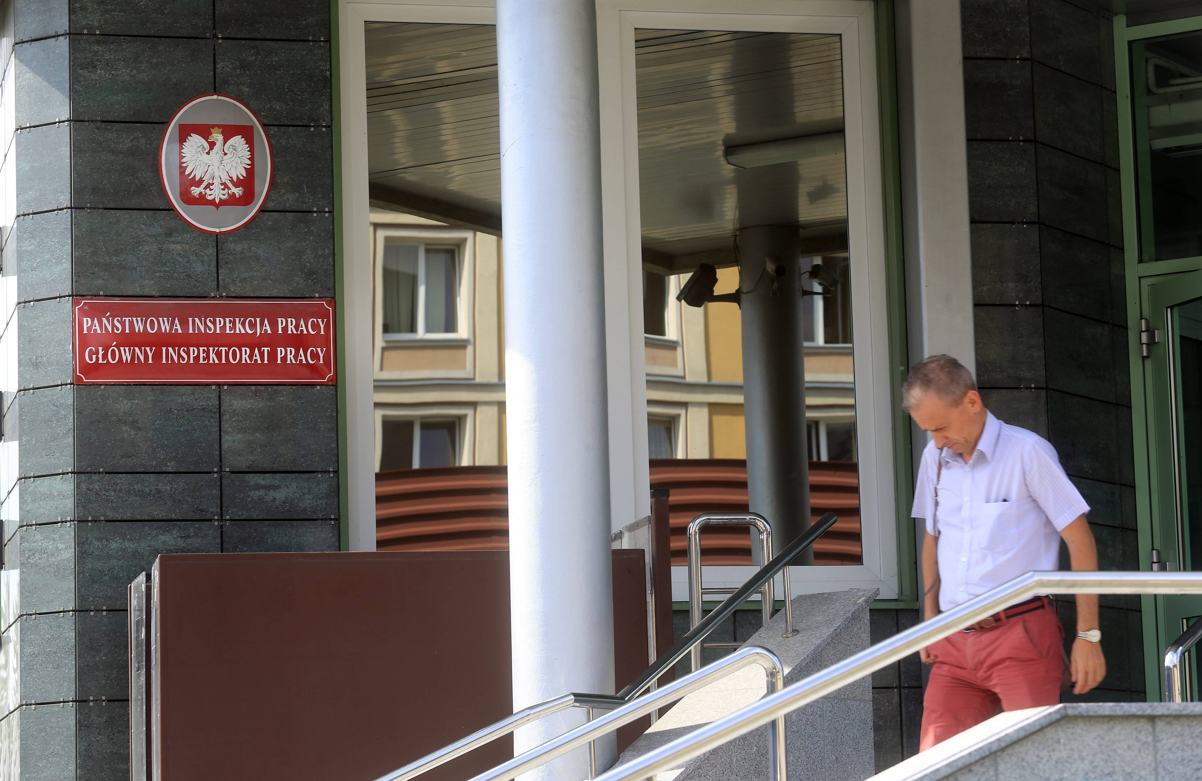 Mężczyzna idzie obok wejścia do budynku, i tablicy informującej, że to Siedziba Państwowej IOnspoekcji Pracy