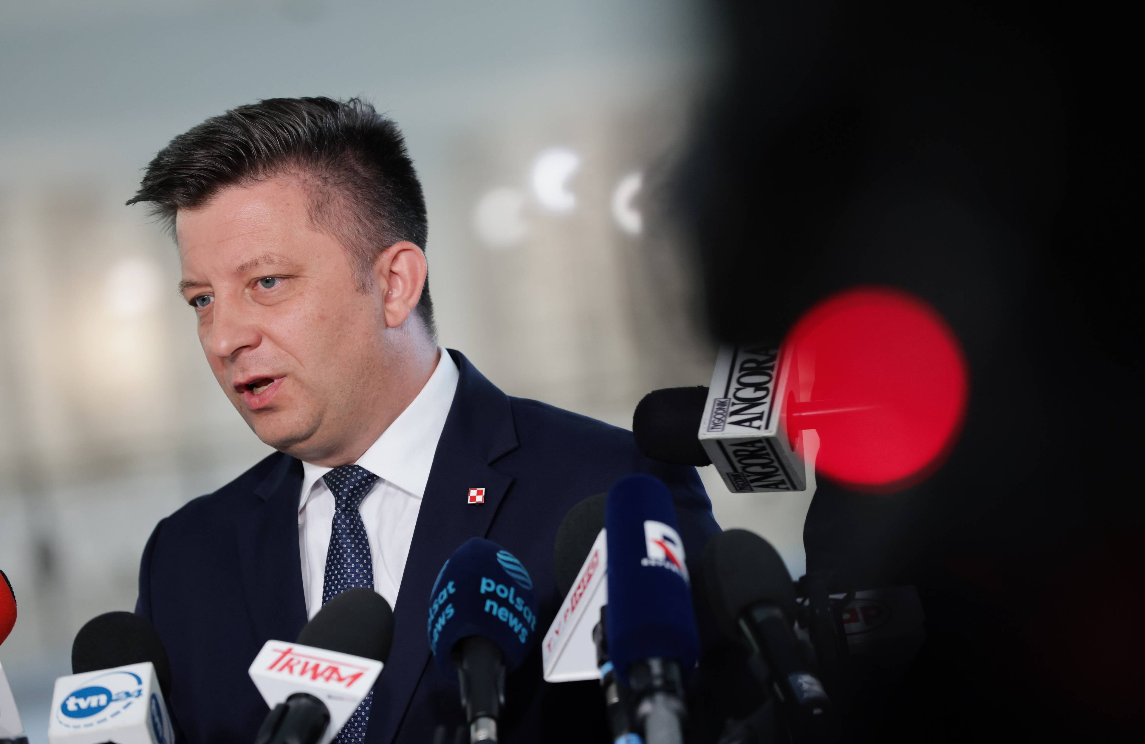 Michał Dworczyk w trakcie konferencji prasowej. Mężczyzna w średnim wieku, w ciemnym garniturze i białej koszuli, stoi przed mikrofonami.