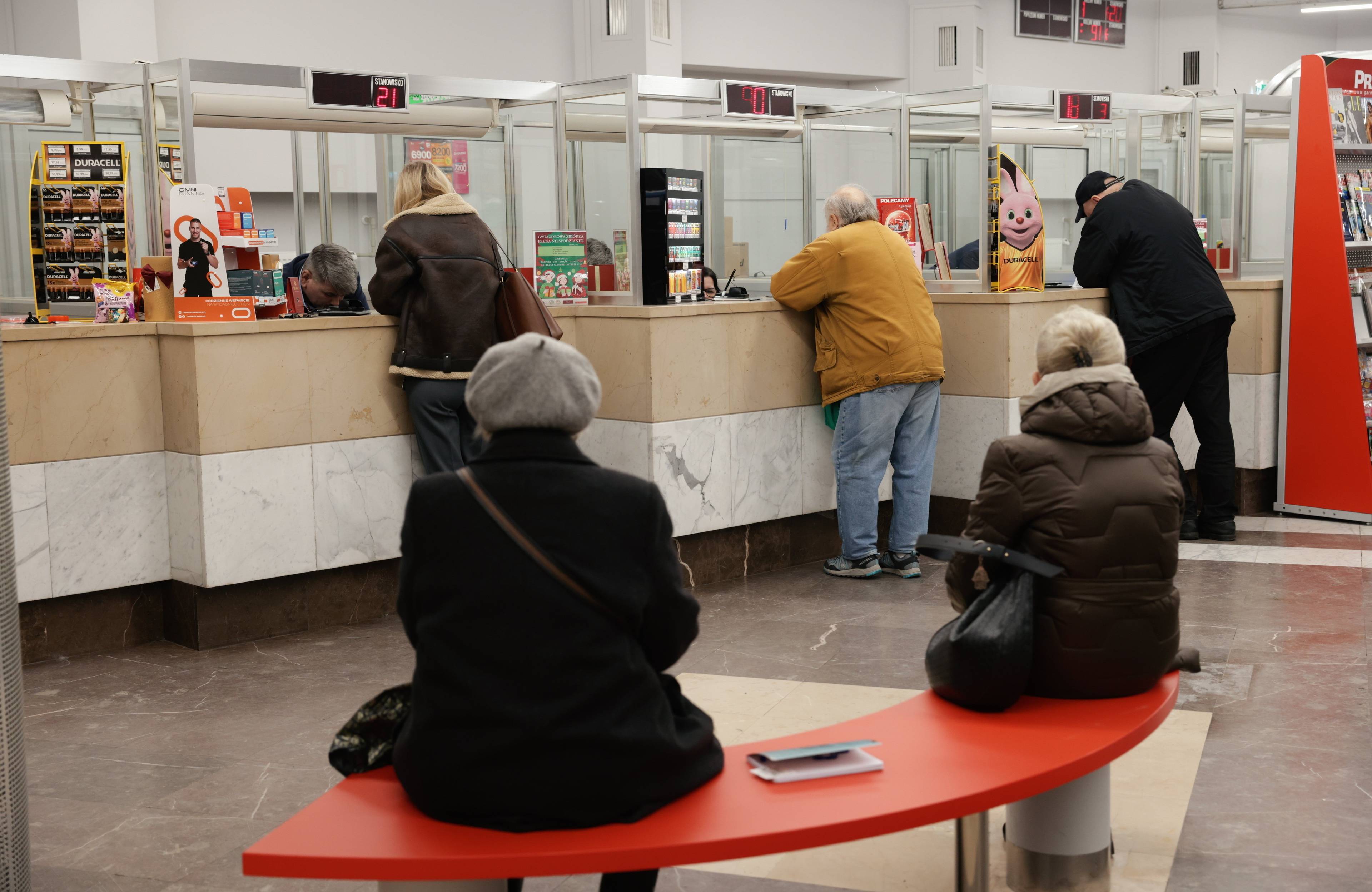 placówka Poczty Polskiej w godzinach pracy, dwie oczekujące osoby na ławce i trzy osoby przy okienkach