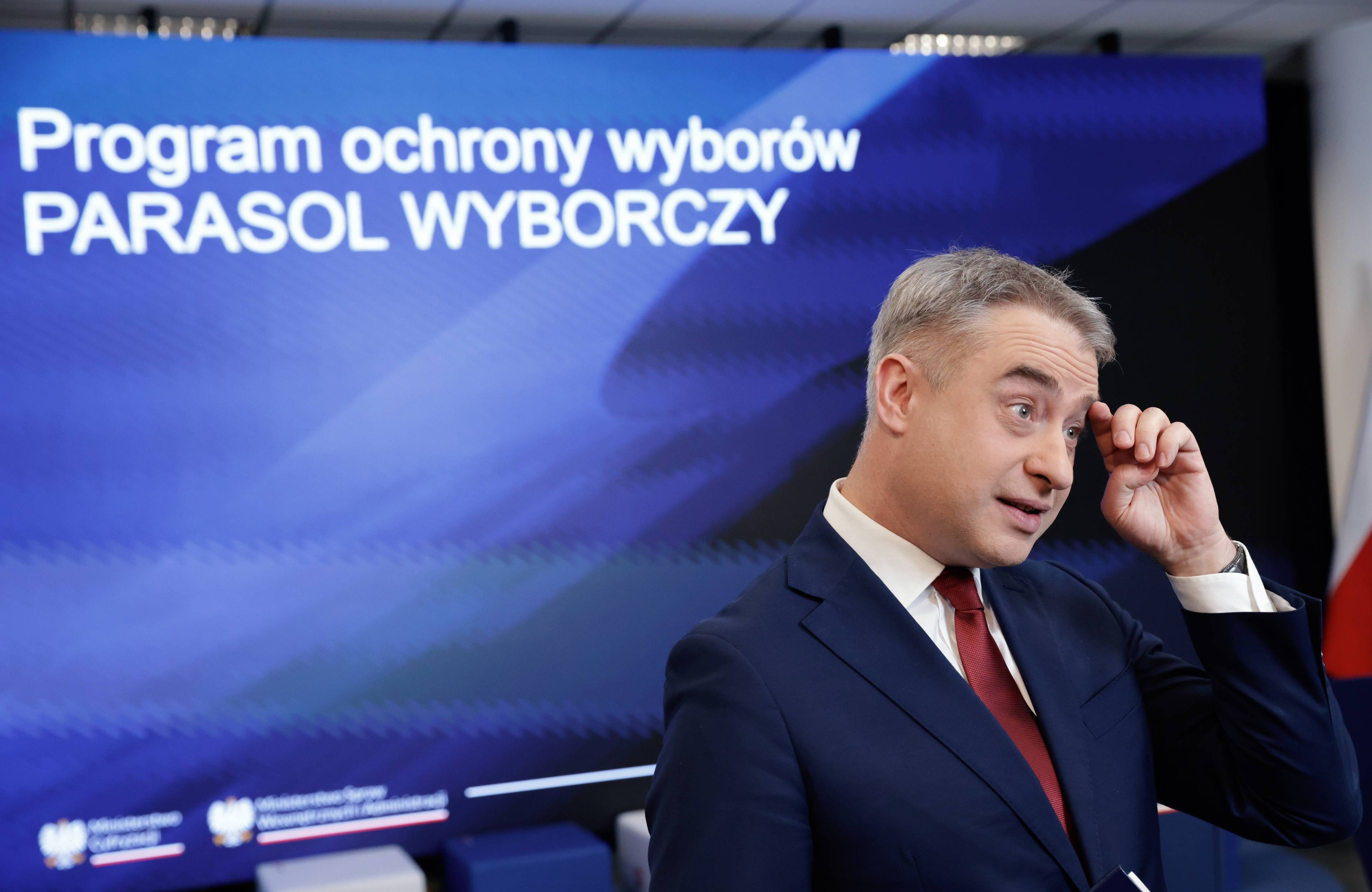 Mężczyzna w ciemnym garniturze i czerwonym krawacie (minister Krzysztof Gawkowski) na tle niebieskiego ekranu z napisem "Program ochrony wyborów PARASOL WYBORCZY"