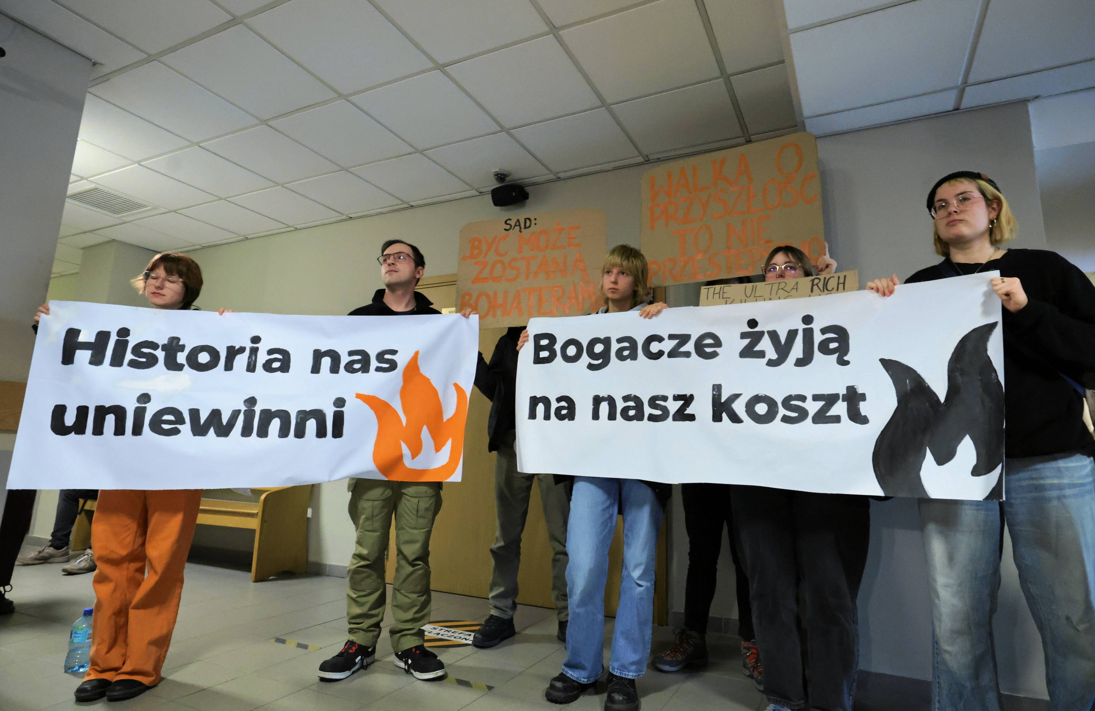 Młodzi ludzie stoją w sądzie z transparentami "Historia nas uniewinni" i "Bogacze żyją na nasz koszt"