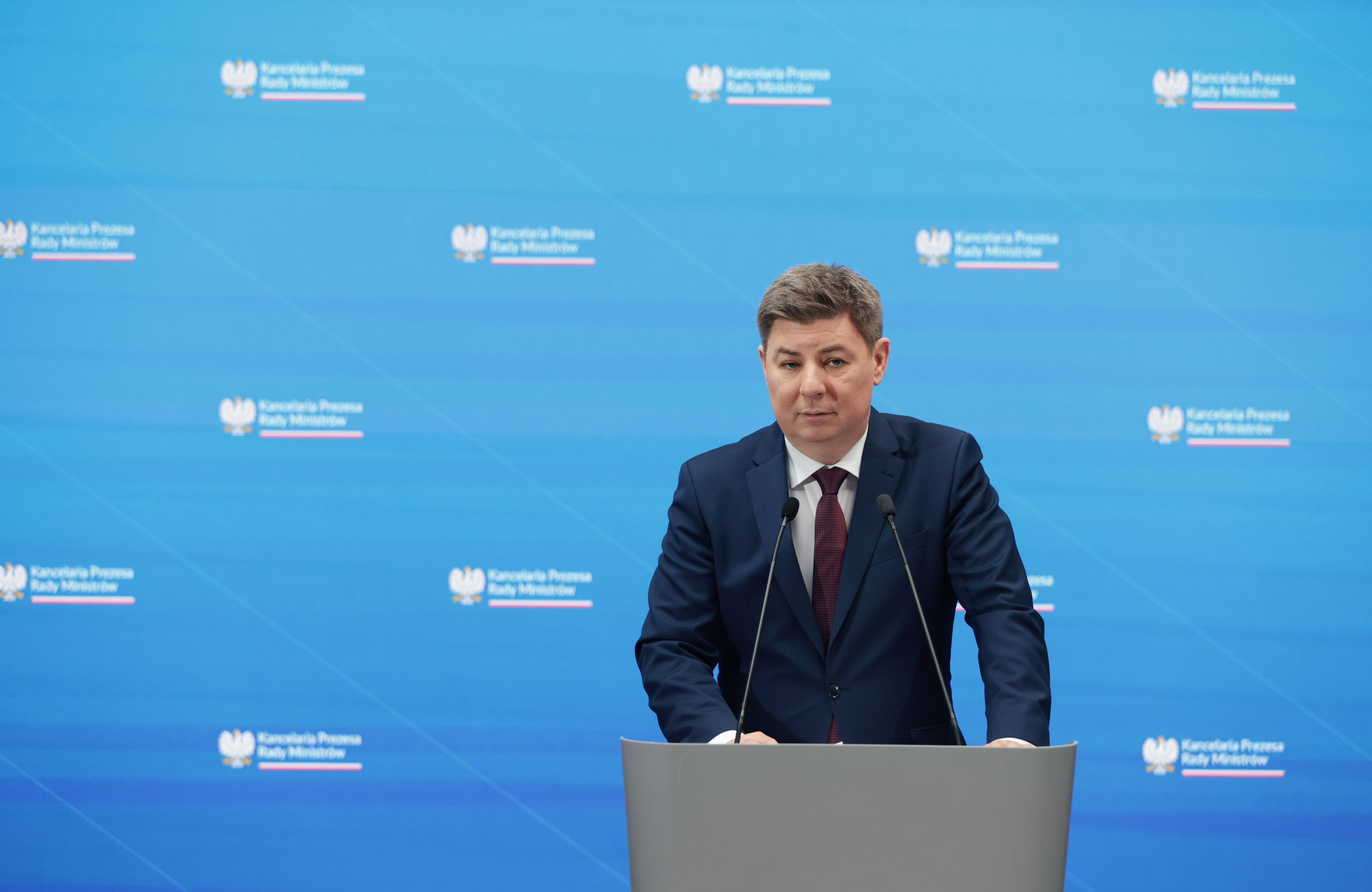Mężczyzna w granatowym garniturze i bordowym krawacie stoi za podium z dwoma mikrofonami na tle niebieskiej ściany z logiem Kancelarii Prezesa Rady Ministrów