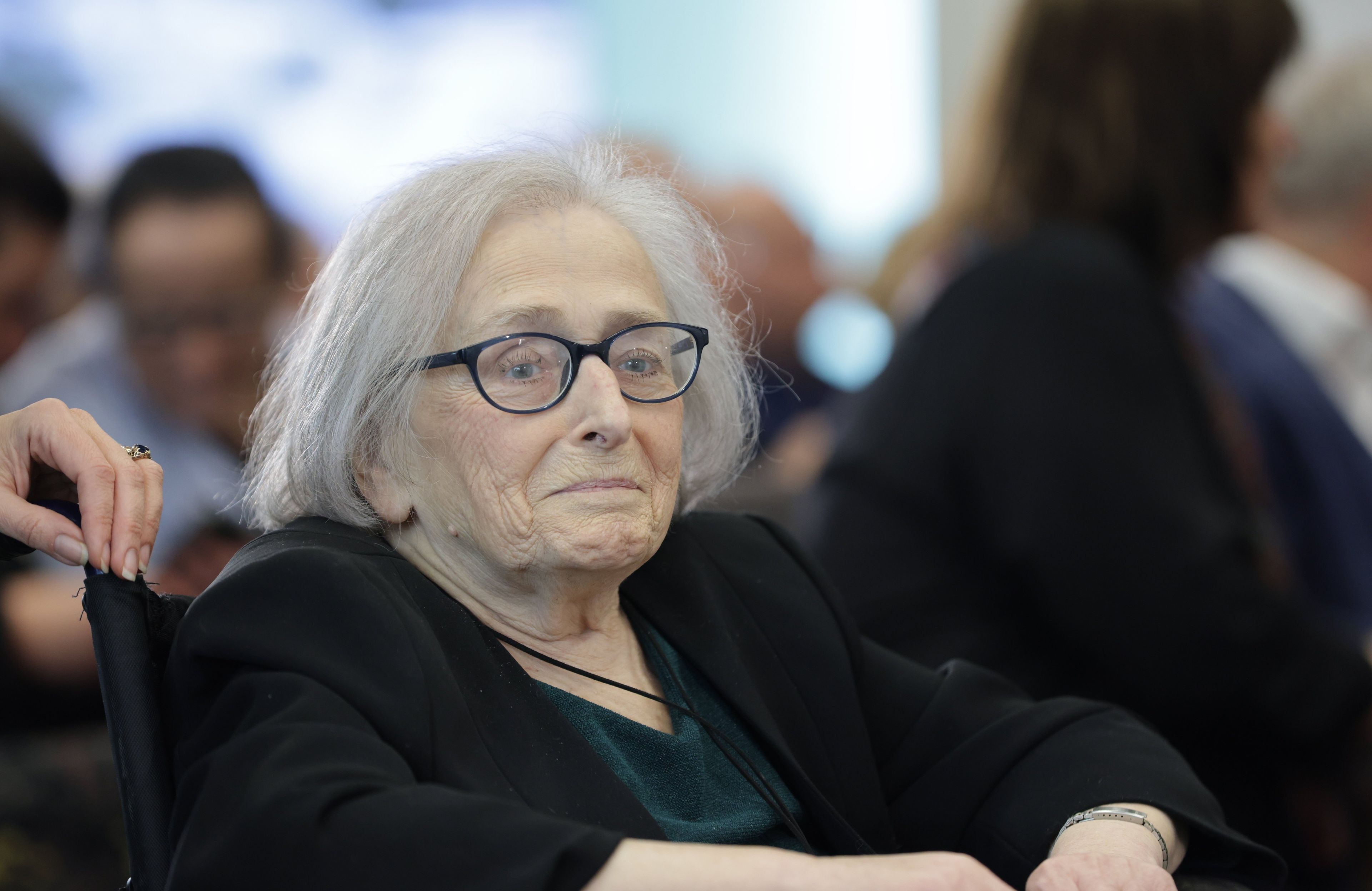 Starsza kobieta o szczupłej twarzy, w czarnych, prostokątnych okularach, o siwych włosach sięgających poniżej ucha, siedzi na wózku inwalidzkim podczas uroczystości zorganizowanej na cześć jej męża. Na jej twarzy widać lekki uśmiech.