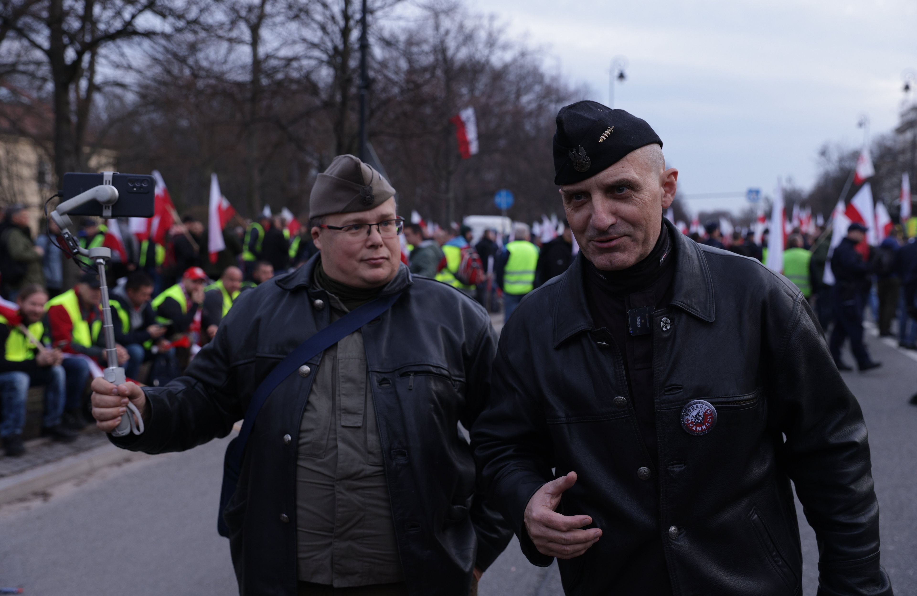 Dwóch mężczyzn w bluzach mundurowych i furażerkach na tle protestujących osób