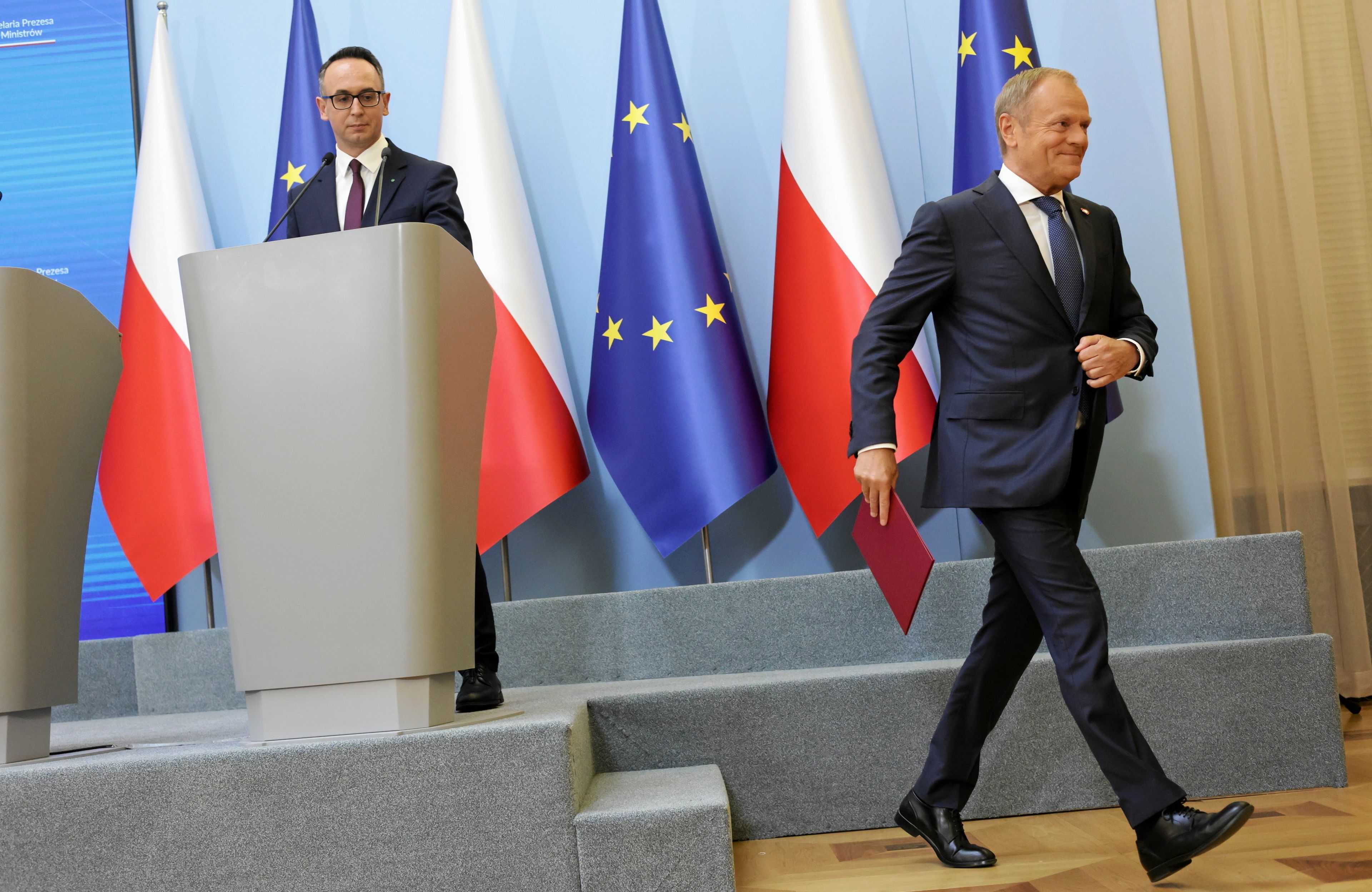 Donald Tusk idzie korytarzem po konferencji na temat powstania CPK, na drugim planie minister infrasktruktury Dariusz Klimczak, flagi polskie i unijne