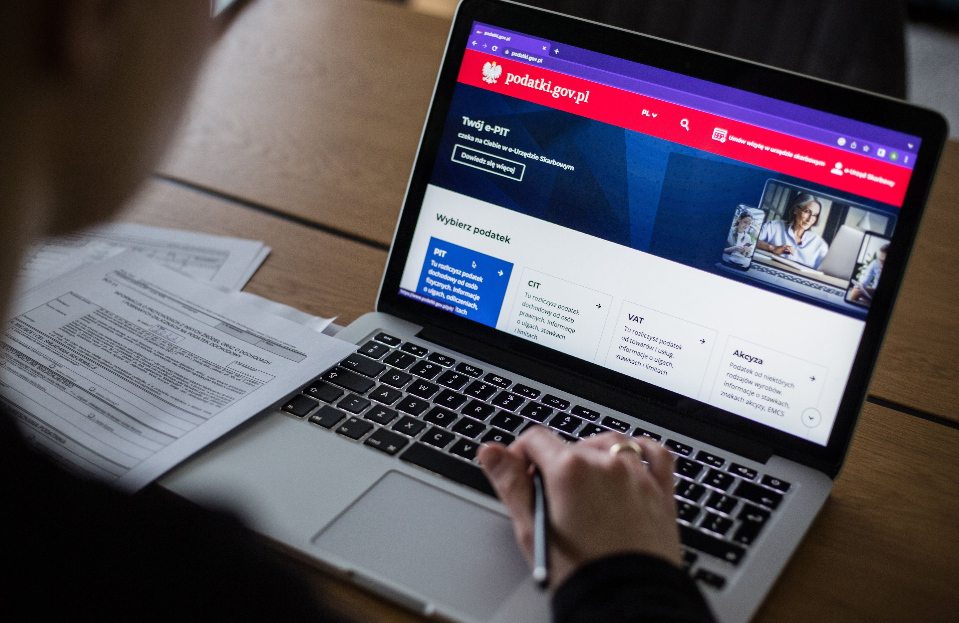 Osoba siedząca przed laptopem, na którym wyświetla się rządowy portal podatki.gov.pl. Obok komputera rozłożone szare druki deklaracji PIT