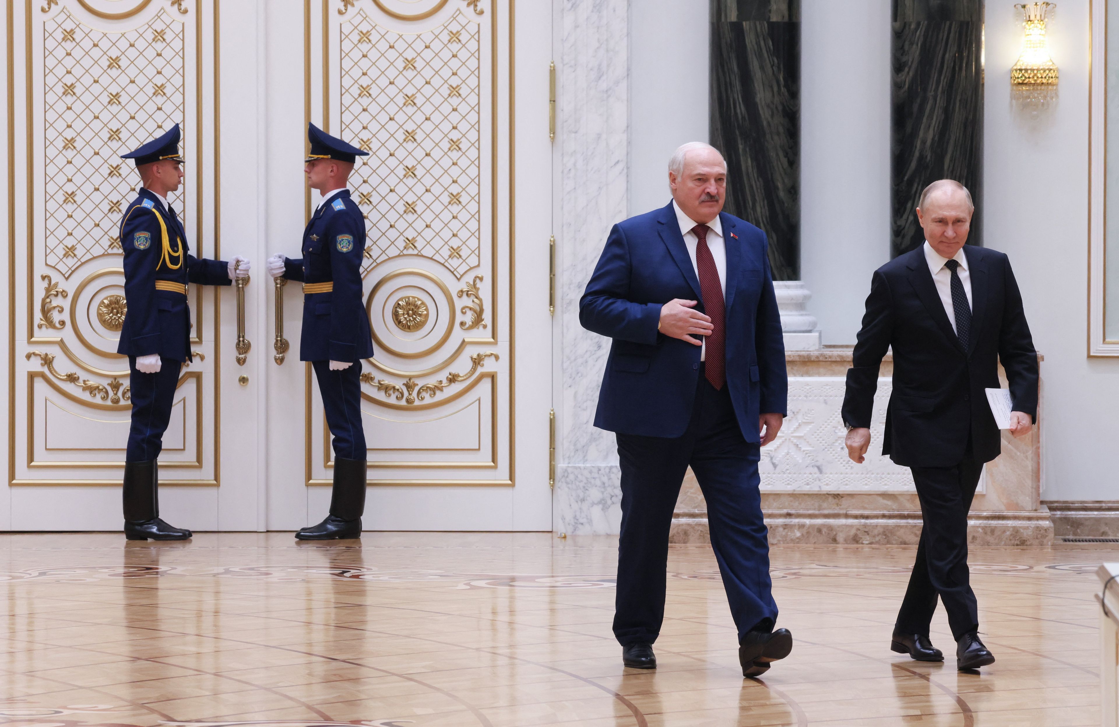 Dwaj mężczyźni w garniturach idą przez salę urządzoną z kiczowatym przepychem.