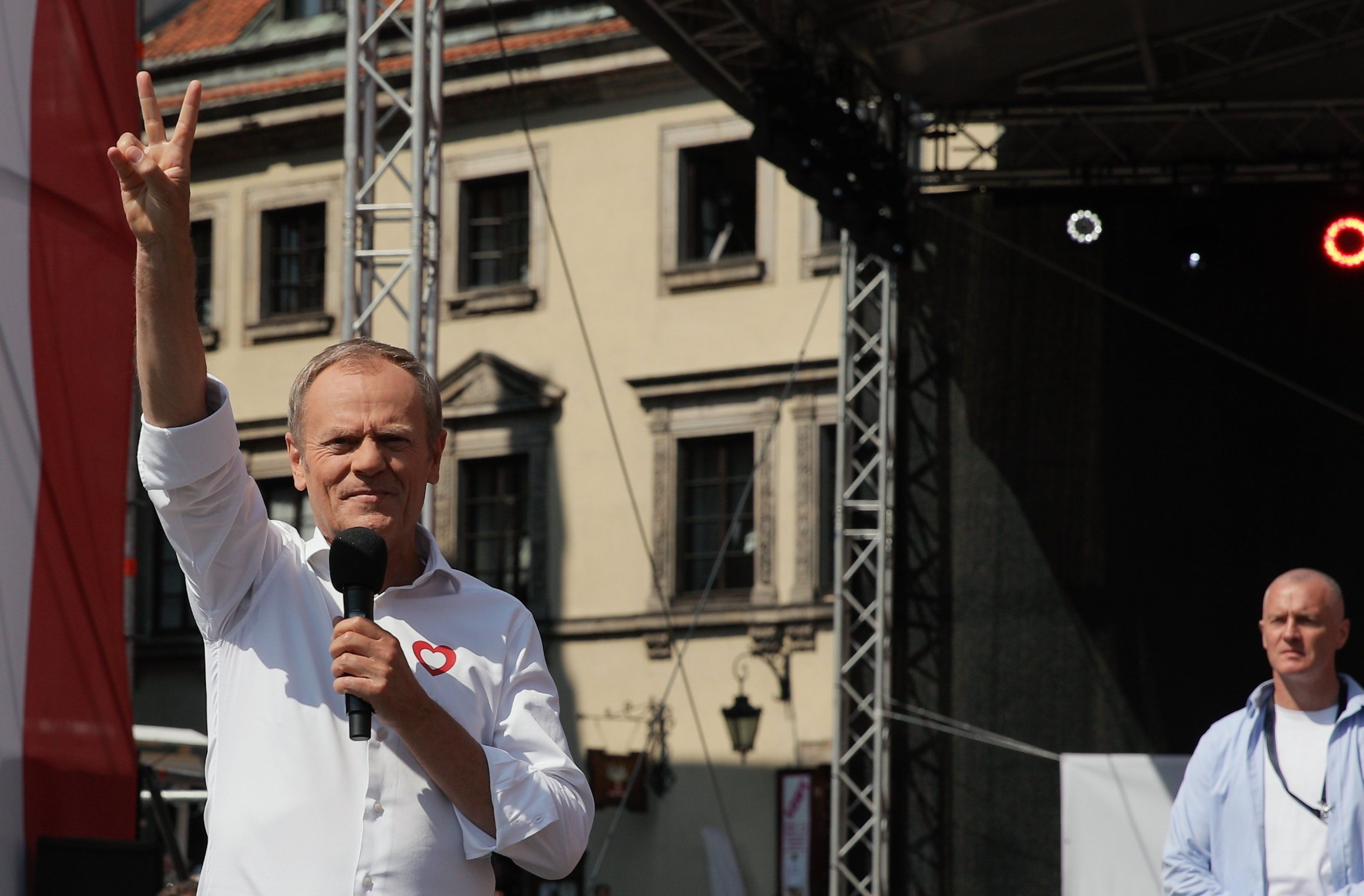 Mężczyzna stoi na scenie, w jednej dłoni trzyma mikrofon, drugą pokazuje znak zwycięstwa