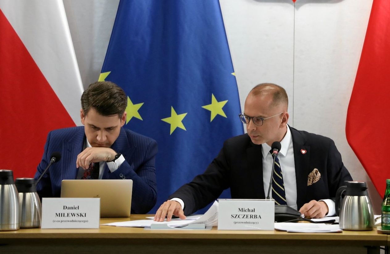 Przewodniczący komisji ds. afery wizowej w Sejmie na tle flag UE i Polski. W środku przewodniczący Michał Szczerba, nad nim godło Polski.