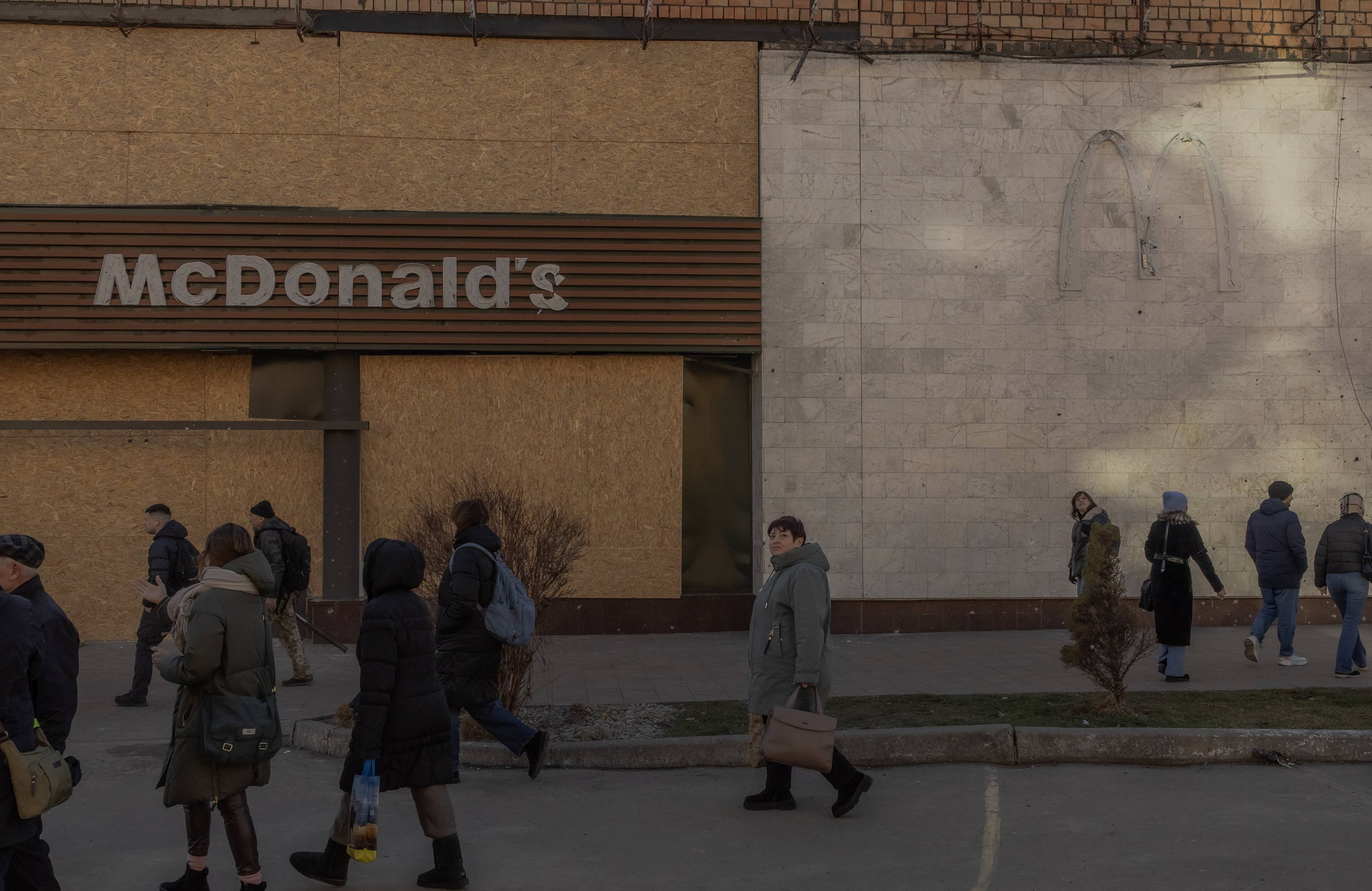 Na zdjęciu widać kilka osób, które przechodzą obok budynku z tablicą Mcdonalda, który został uszkodzony. Jego wybite szyby zostały zastawione dyktą