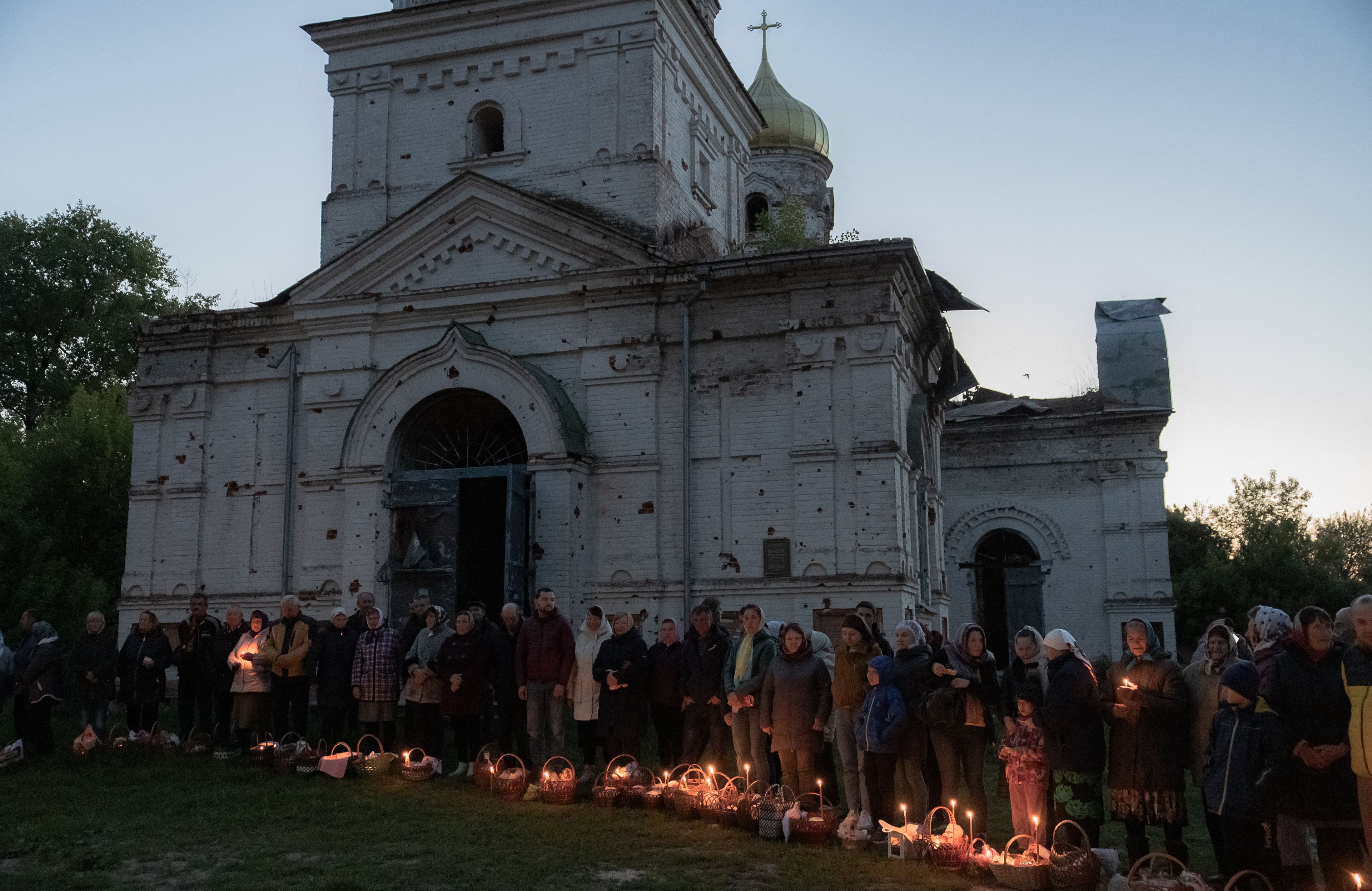 Świt. Na zdjęciu są ludzie, święcą pokarmy obok zniszczonej cerkwi. W koszyczkach palą się świeczki. Wielkanoc prawosławna