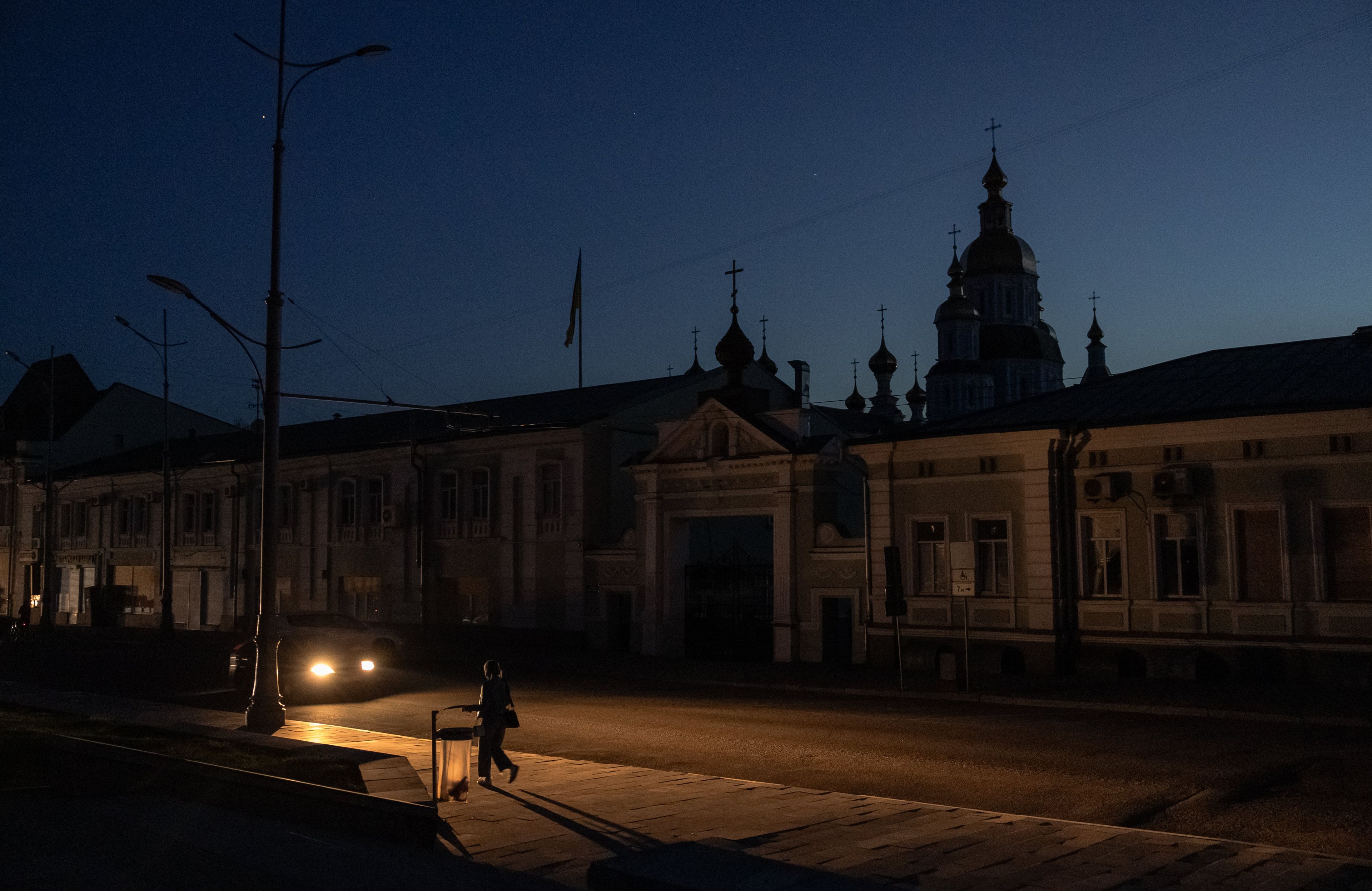 Na zdjęciu jest ciemna ulica, po której idzie osoba. Z tyłu są światła samochodu