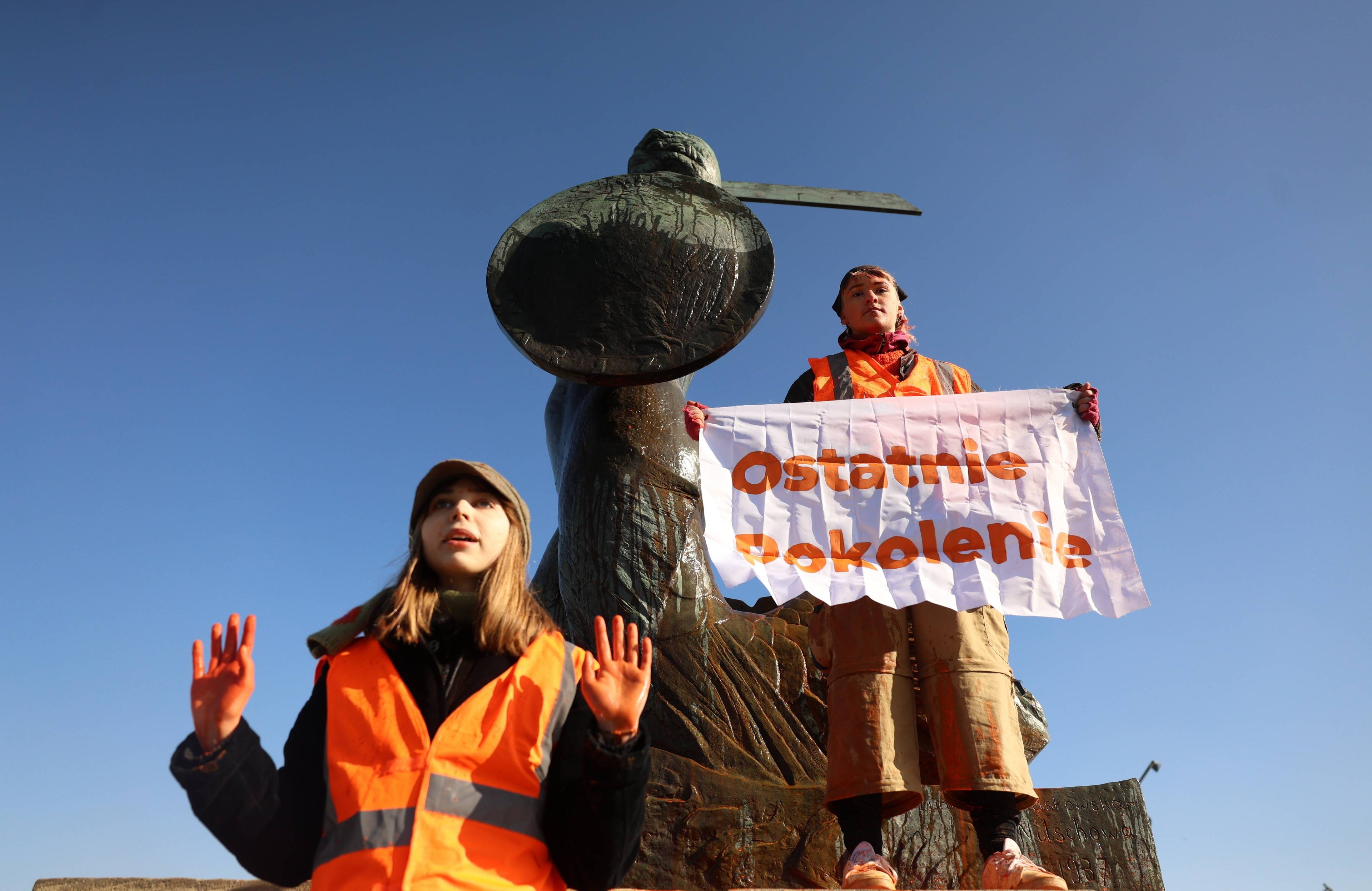 dwójka aktywistów, jedno z nich trzyma napis "ostatnie pokolenie", stoją na pomniku syrenki