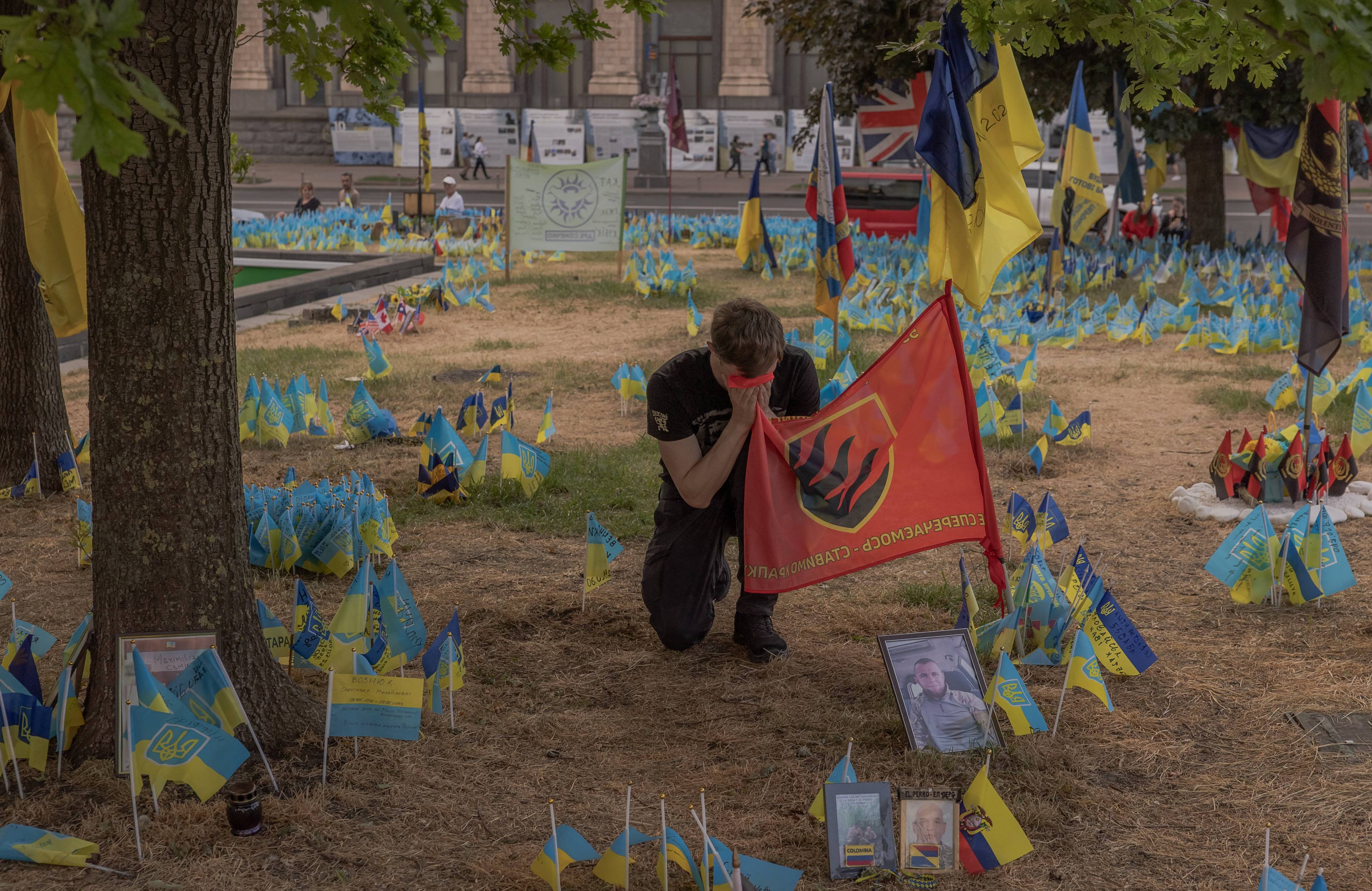 żołnierz klęczy i całuje flagę brygady pośród zatkniętych w ziemię flag ukraińskich