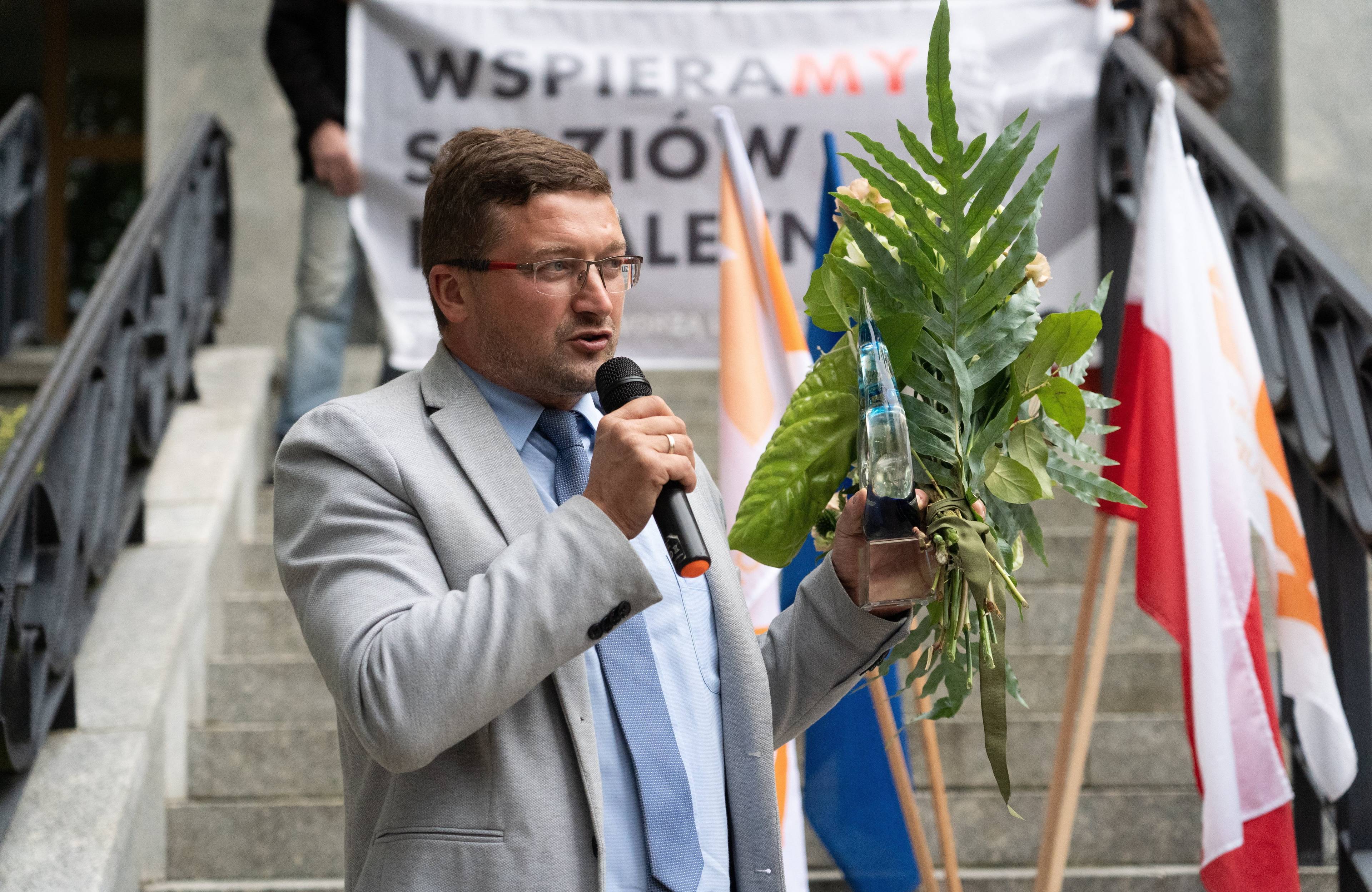 Mężczyzna w szarej marybarce z mikrofonem w ręku przed budynkiem sądu w Olsztynie