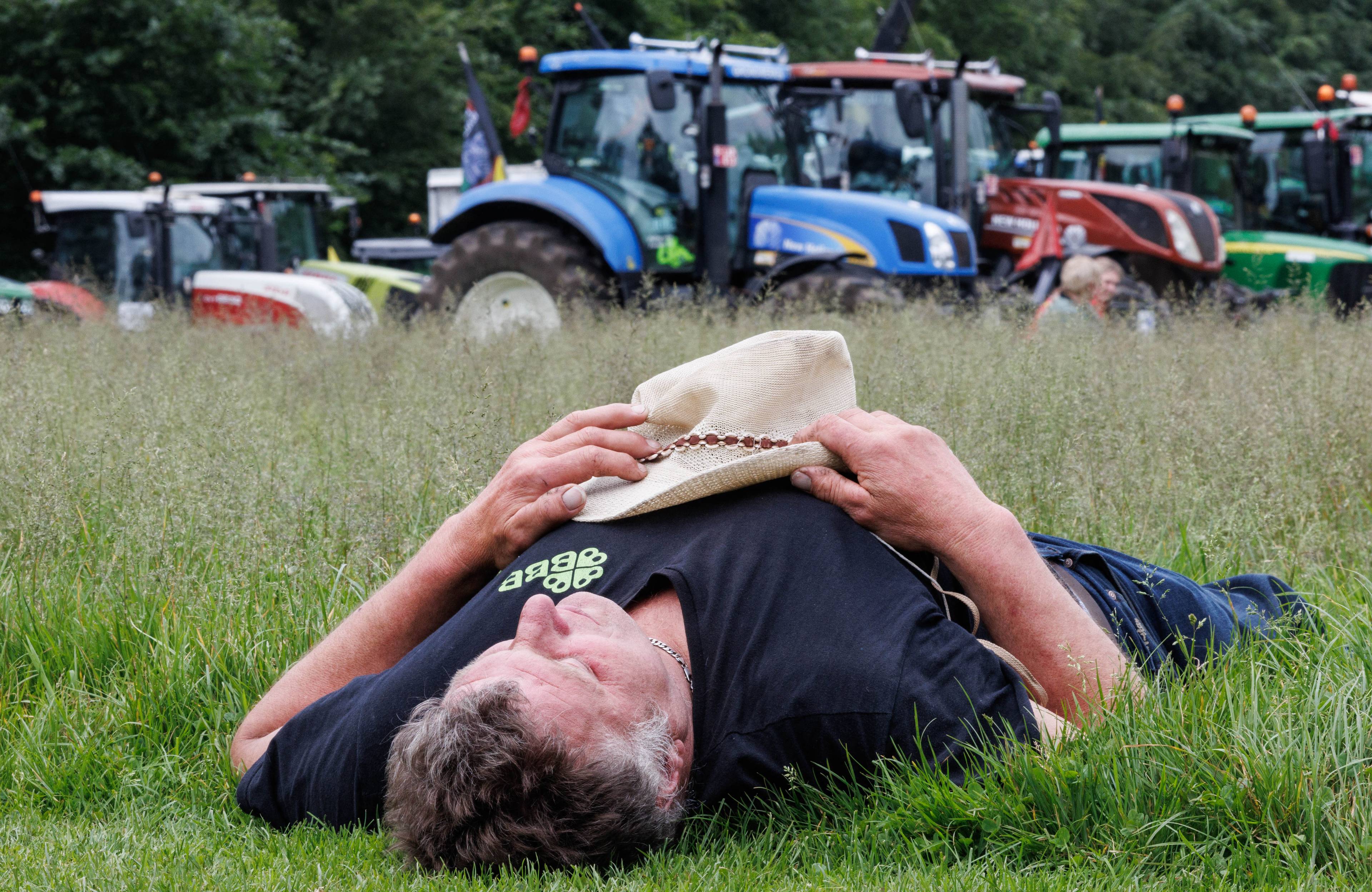 leżący na trawie mężczyzna trzyma na brzuchu kapelusz, w tle traktory