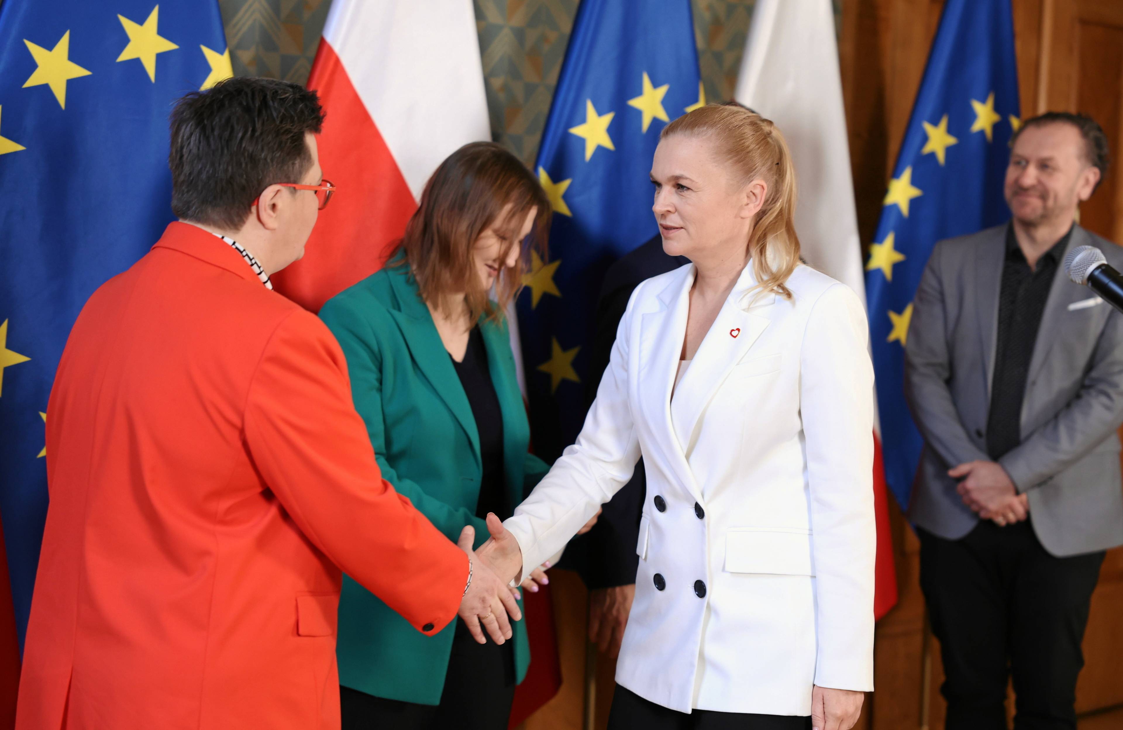Trzy kobiety: w czerwonym, białym i zielonym żakiecie na tle polskich i unijnych flag