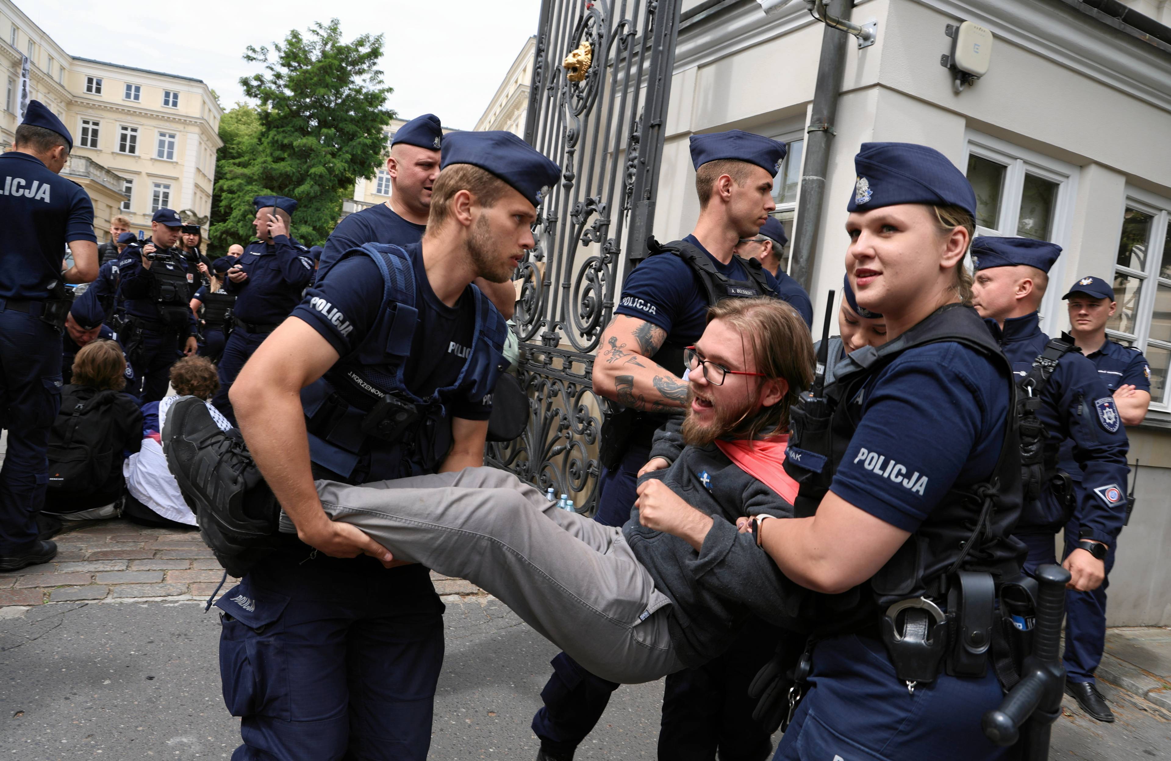 Czterech policjantów w granatowych mundurach wynosi za ręce i nogi młodego brodatego mężczyznęz w okularach ze związanymi blond włosami