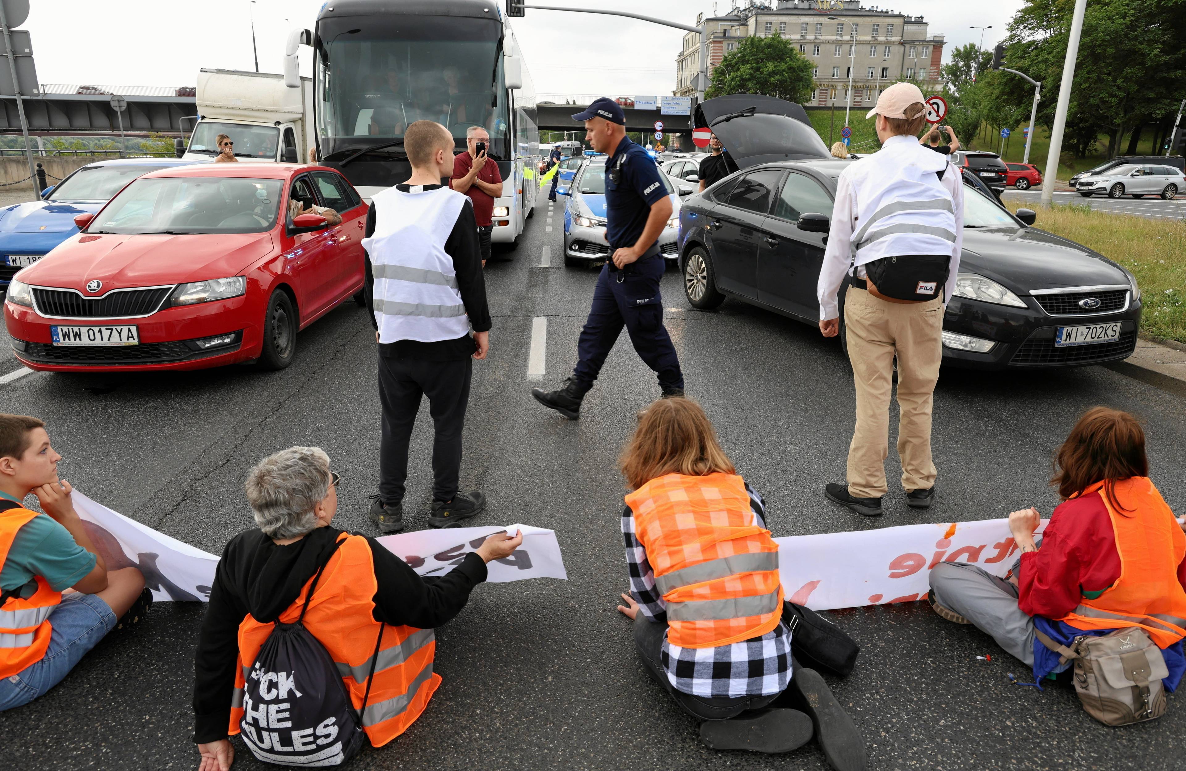 aktywiści siedzą na jezdni w kamizelkach ostrzegawczych, blokują jezdnię, na zdjęciu widać też policję